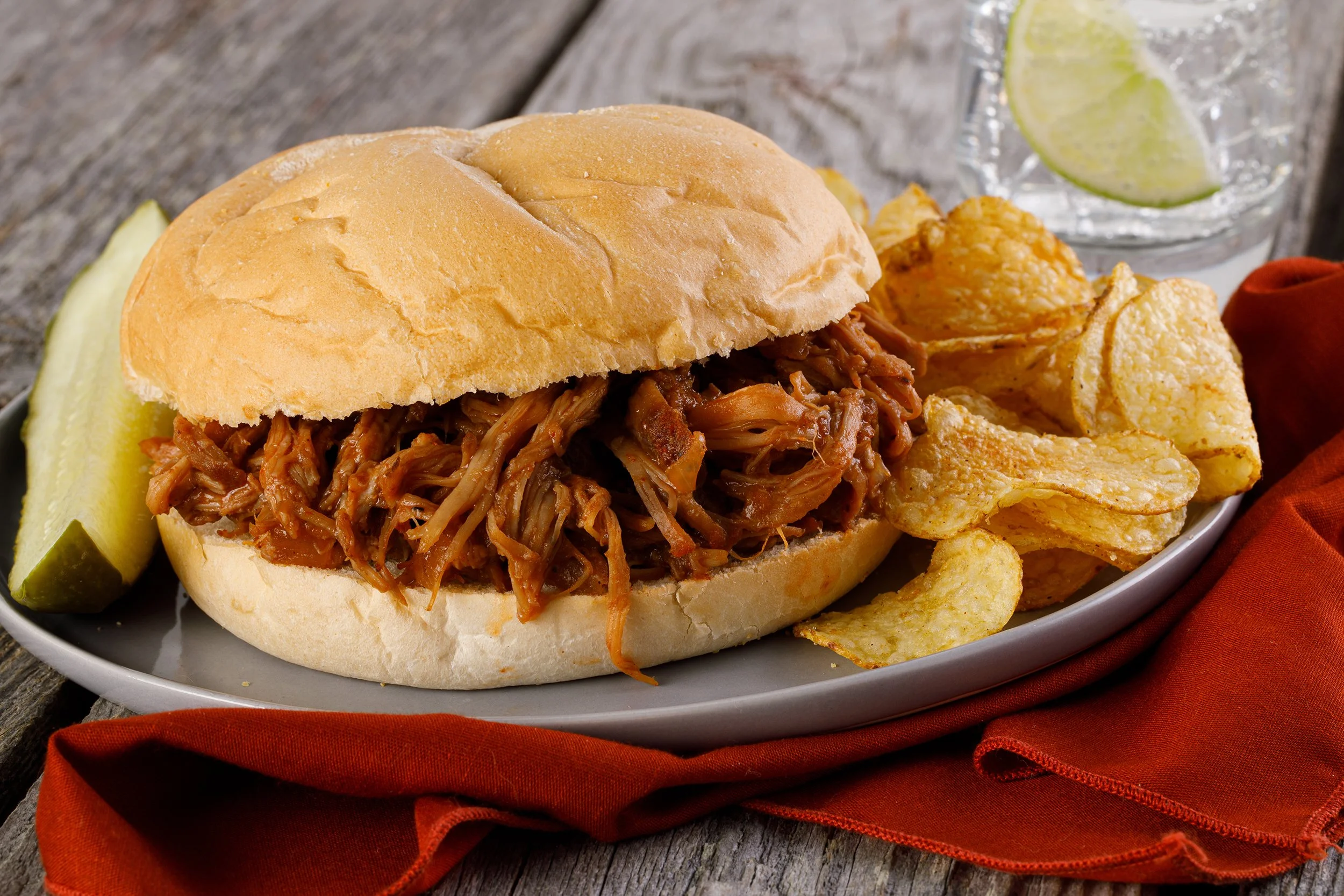 Pulled pork sandwich with potato chips, a pickle, and a glass of lime-infused water on a rustic wooden table.