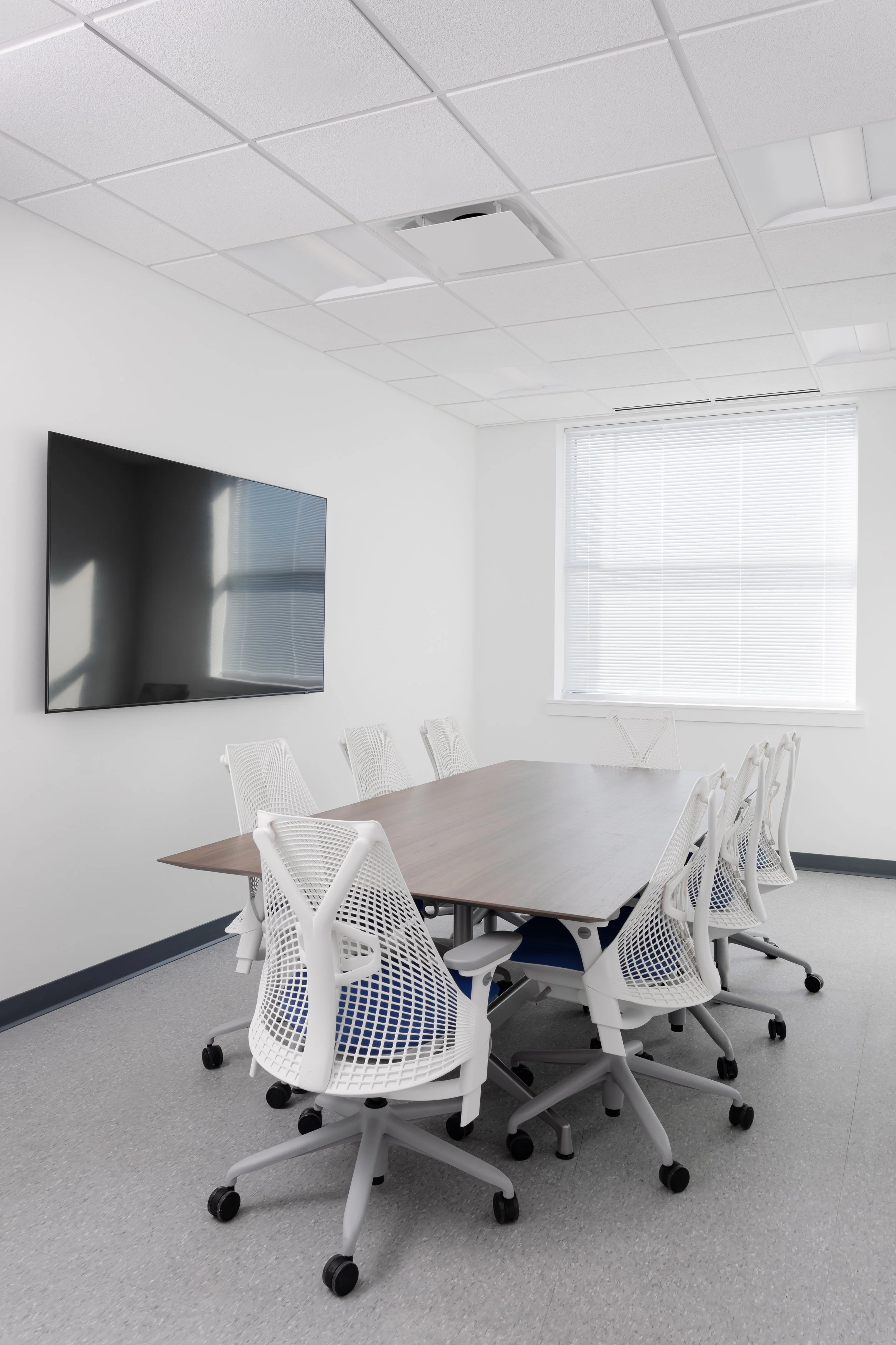 Empty conference room with a rectangular table, white ergonomic chairs on wheels, a wall-mounted flat-screen TV, and a window with blinds.