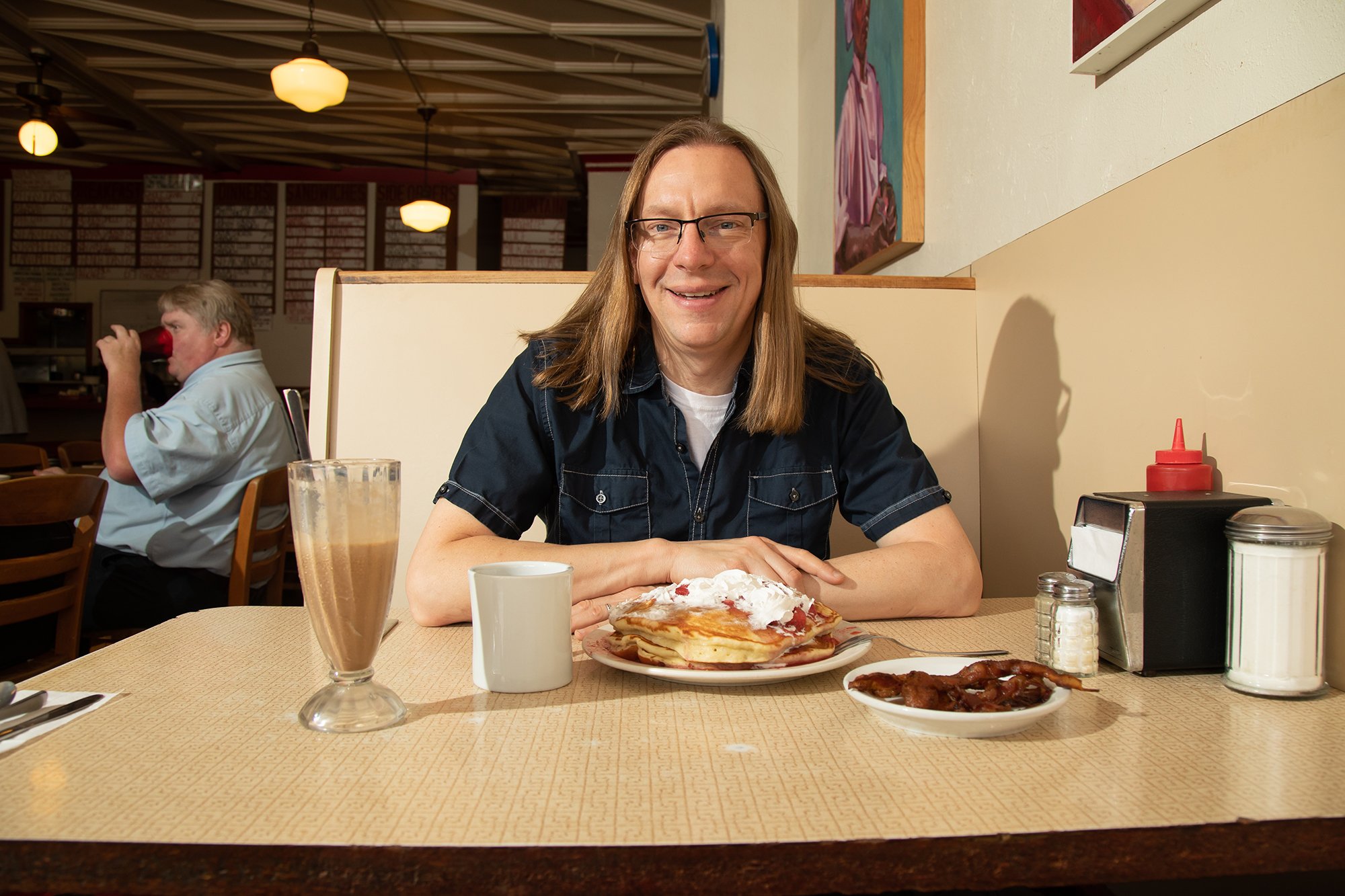 Man with long hair and glasses smiling at the camera in a diner. On the table in front of him is a stack of pancakes with whipped cream and syrup, a latte, a cup of coffee, and a small plate of bacon. The background features another man drinking from