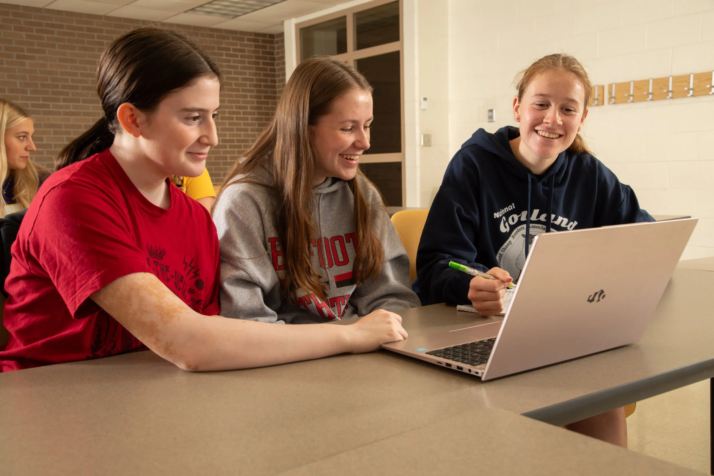 Four young women sitting at a table in a classroom, three of them looking at a laptop computer while smiling. One of them has a pen in hand and appears engaged in the activity.