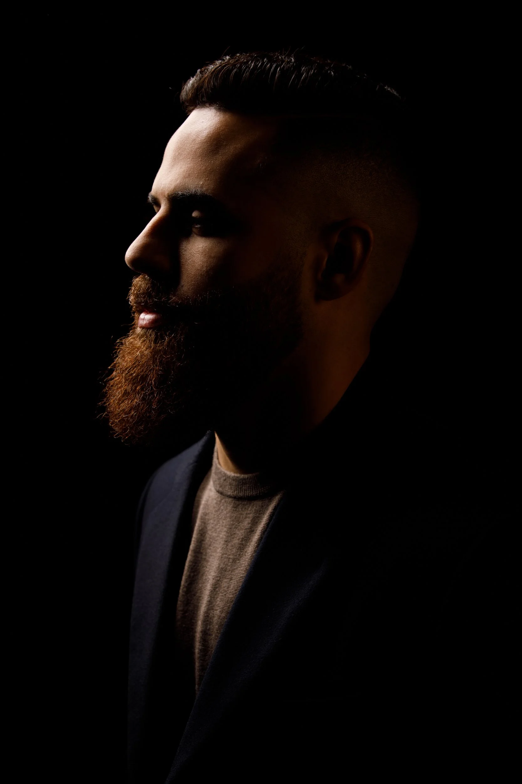 A side profile of a man with a well-groomed beard, styled dark hair, and wearing a blazer over a beige shirt, set against a black background.