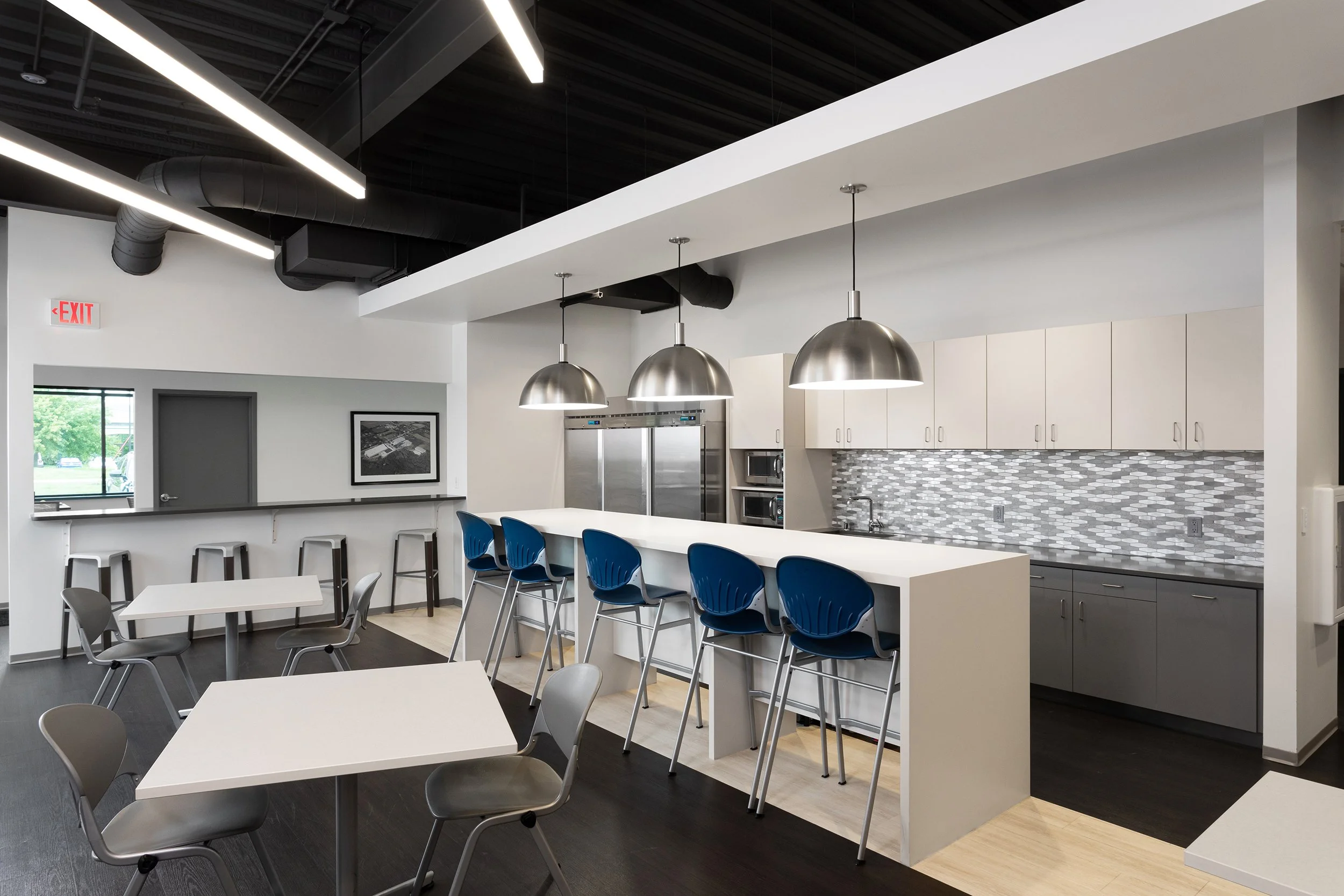 Modern kitchen and dining area with white counters, blue bar stools, gray and white chairs, stainless steel appliances, and pendant lighting.