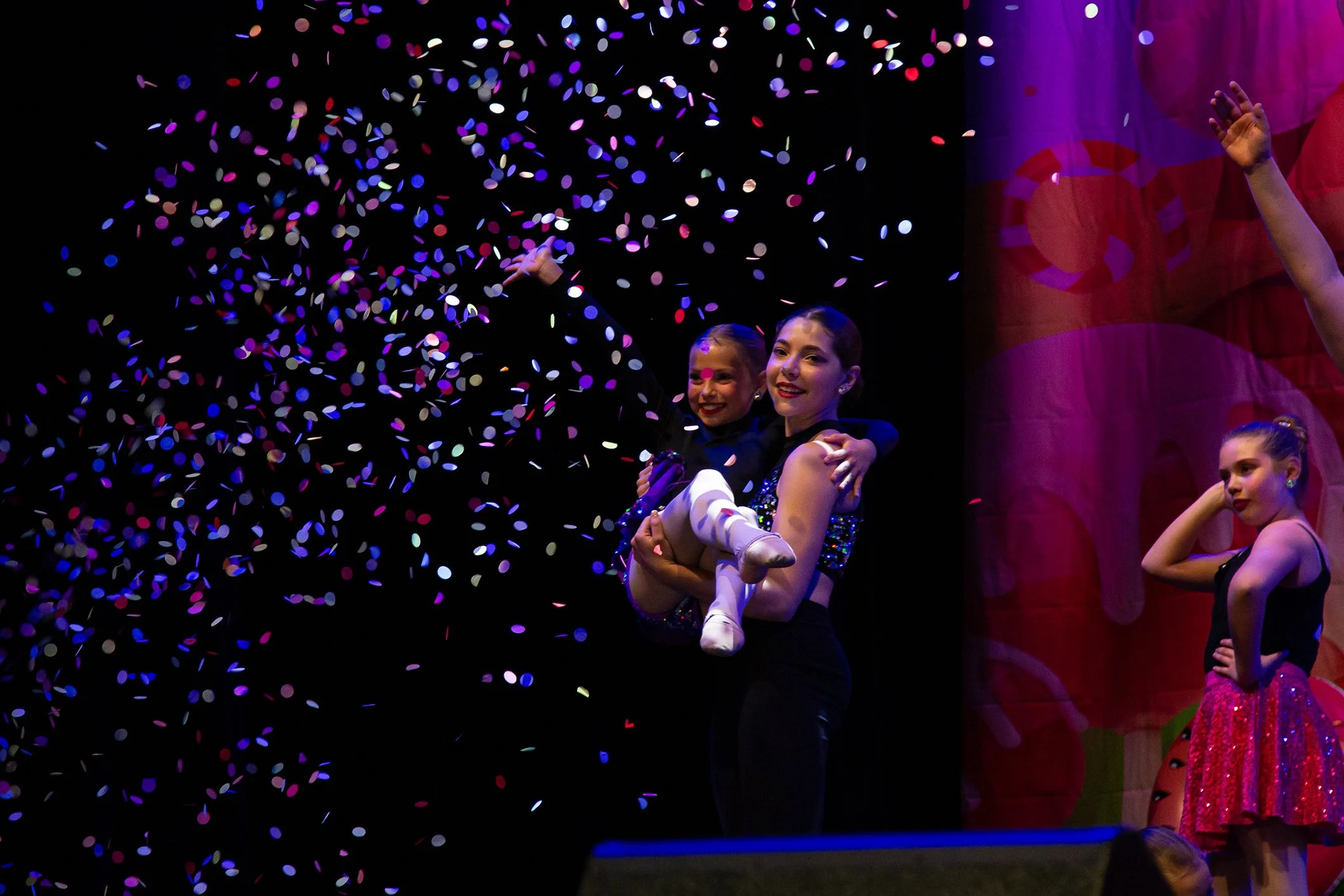 Two young girls, dressed in dance costumes, are on stage during a performance, with one girl being held by an older dancer while colorful confetti falls around them.
