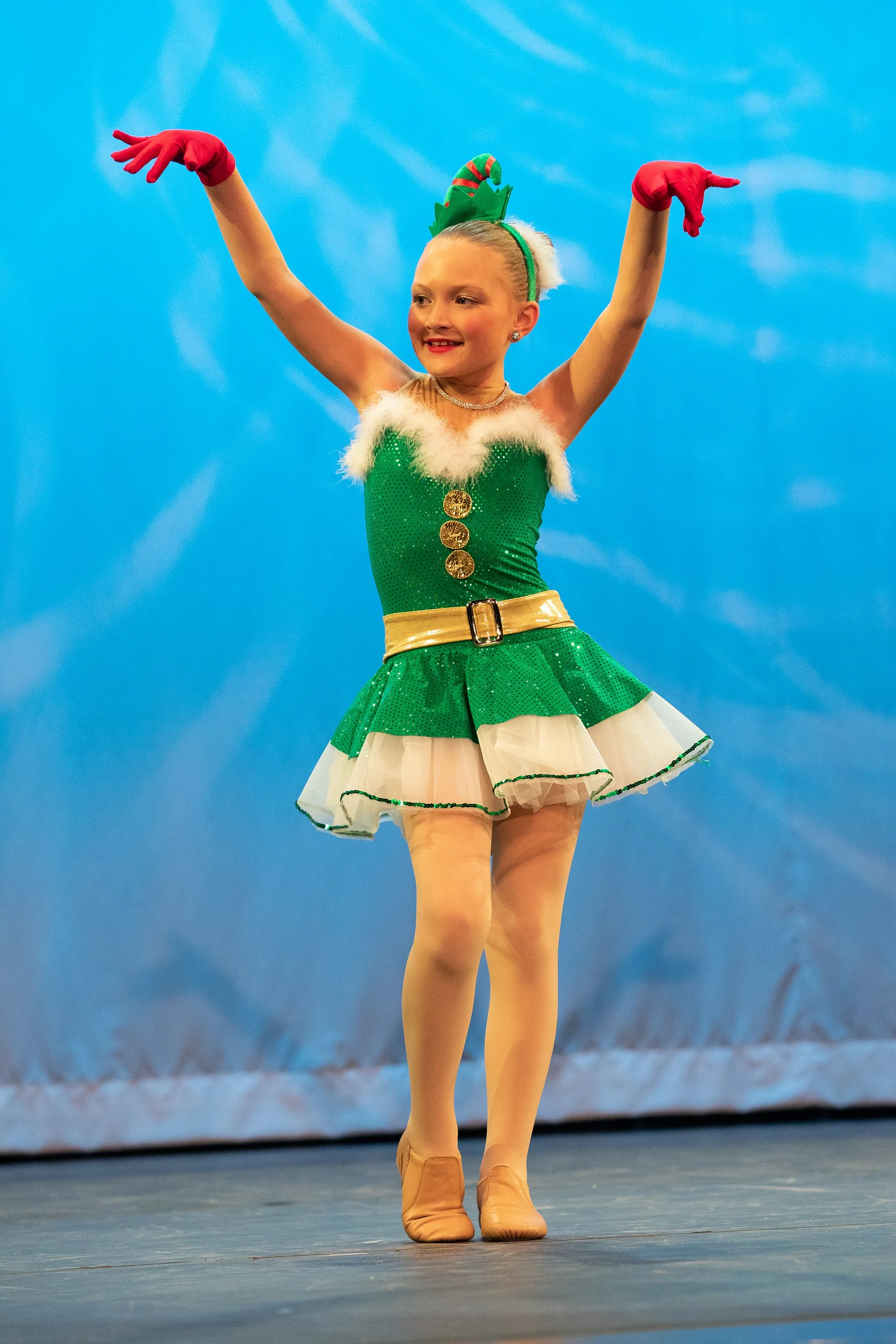 Young girl in a festive green and white costume, performing a dance on stage, with a blue backdrop.