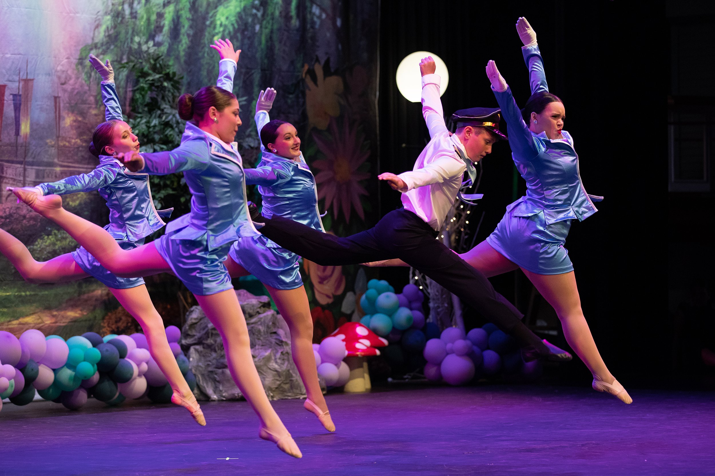 Group of dancers performing a leap on stage, dressed in blue costumes with one dancer in a white shirt and police hat, with a colorful and whimsical backdrop decorated with balloons and large mushrooms.