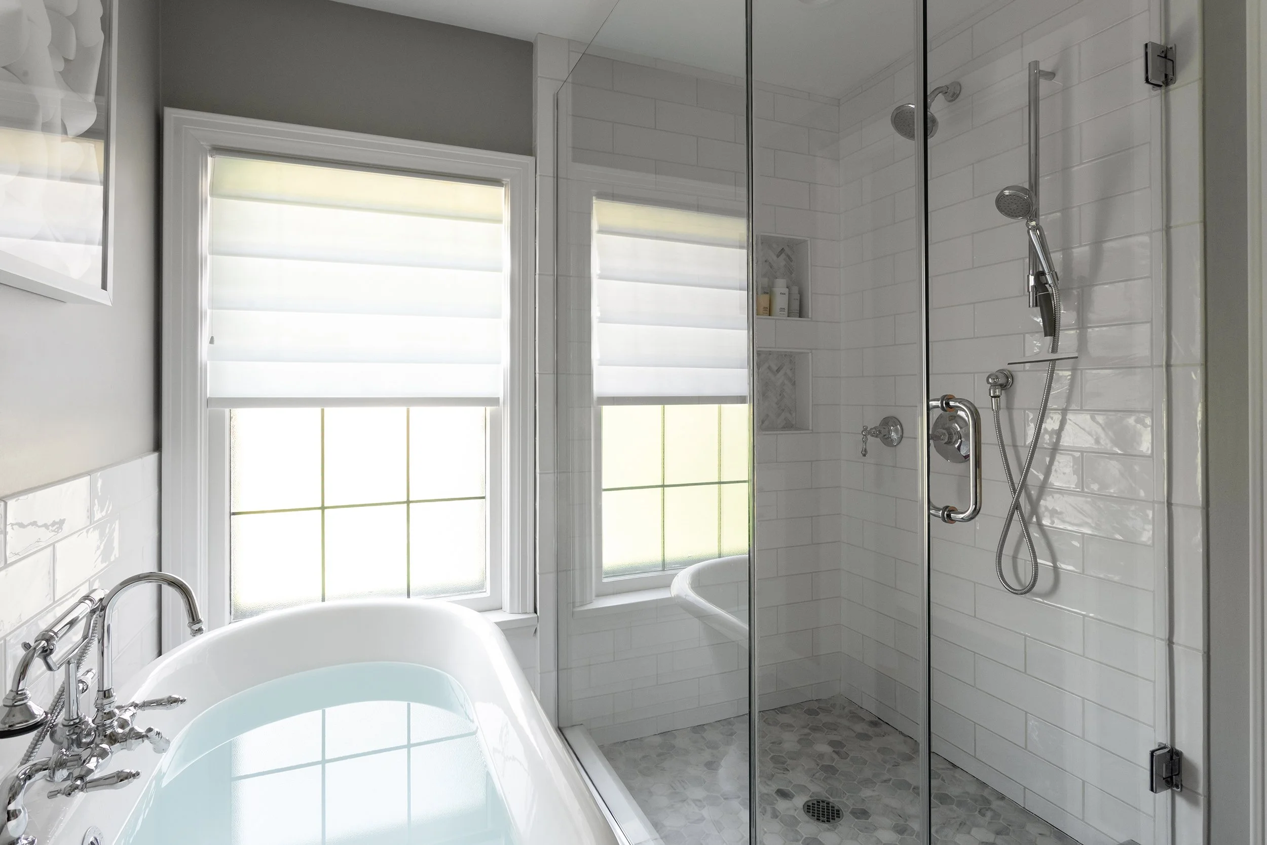 A modern bathroom featuring a bathtub filled with water, a walk-in shower with a glass door, window with shades, white tiled walls, and a small built-in shelf for toiletries.
