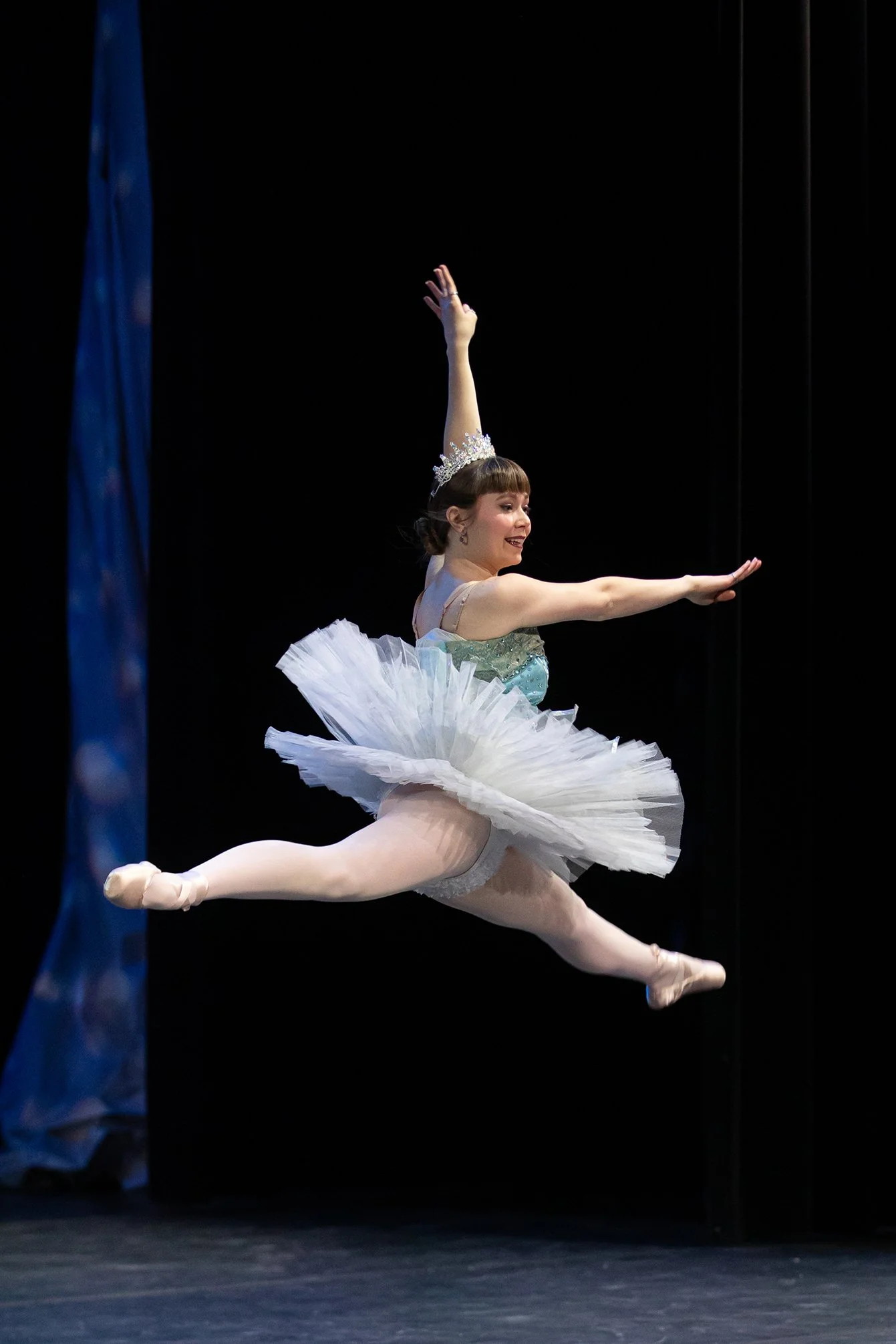A ballet dancer in a tutu and tiara performing a leap on stage.