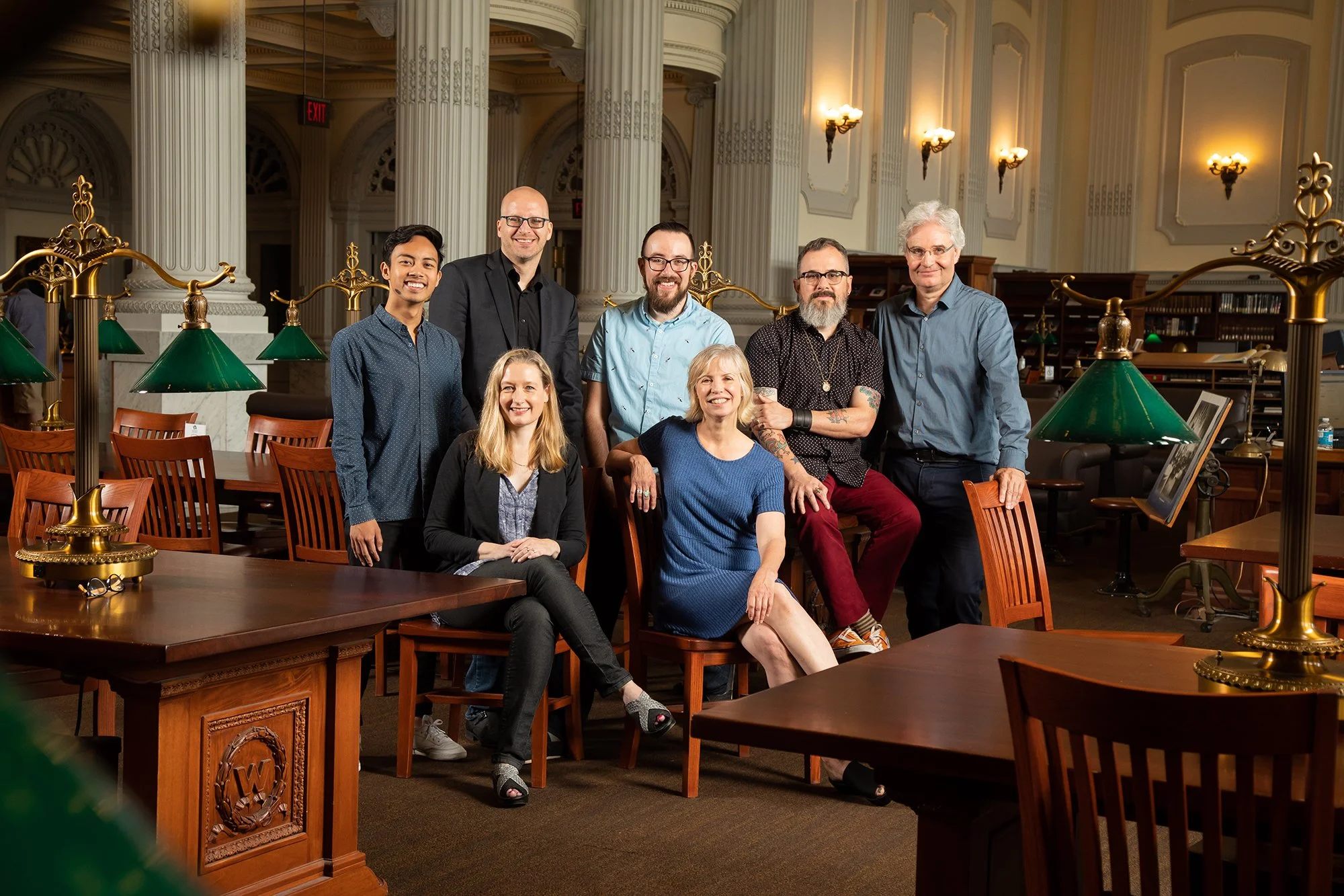 Group of eight people posing inside a library with tall classical columns, ornate wall sconces, and wooden bookshelves in the background.