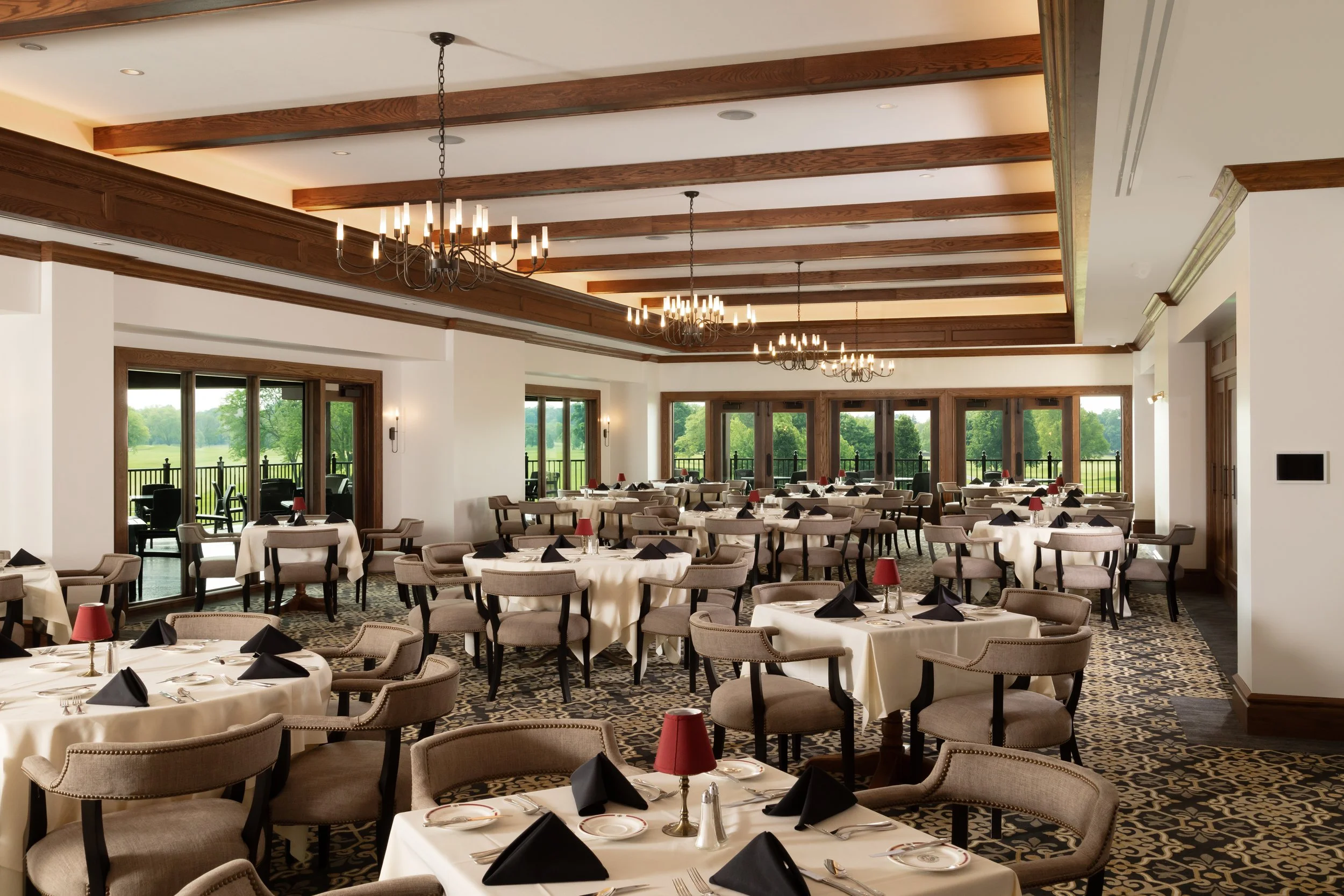 Elegant restaurant with round tables covered in white tablecloths, set with black napkins, utensils, and red lamps, spacious with large windows and chandeliers.