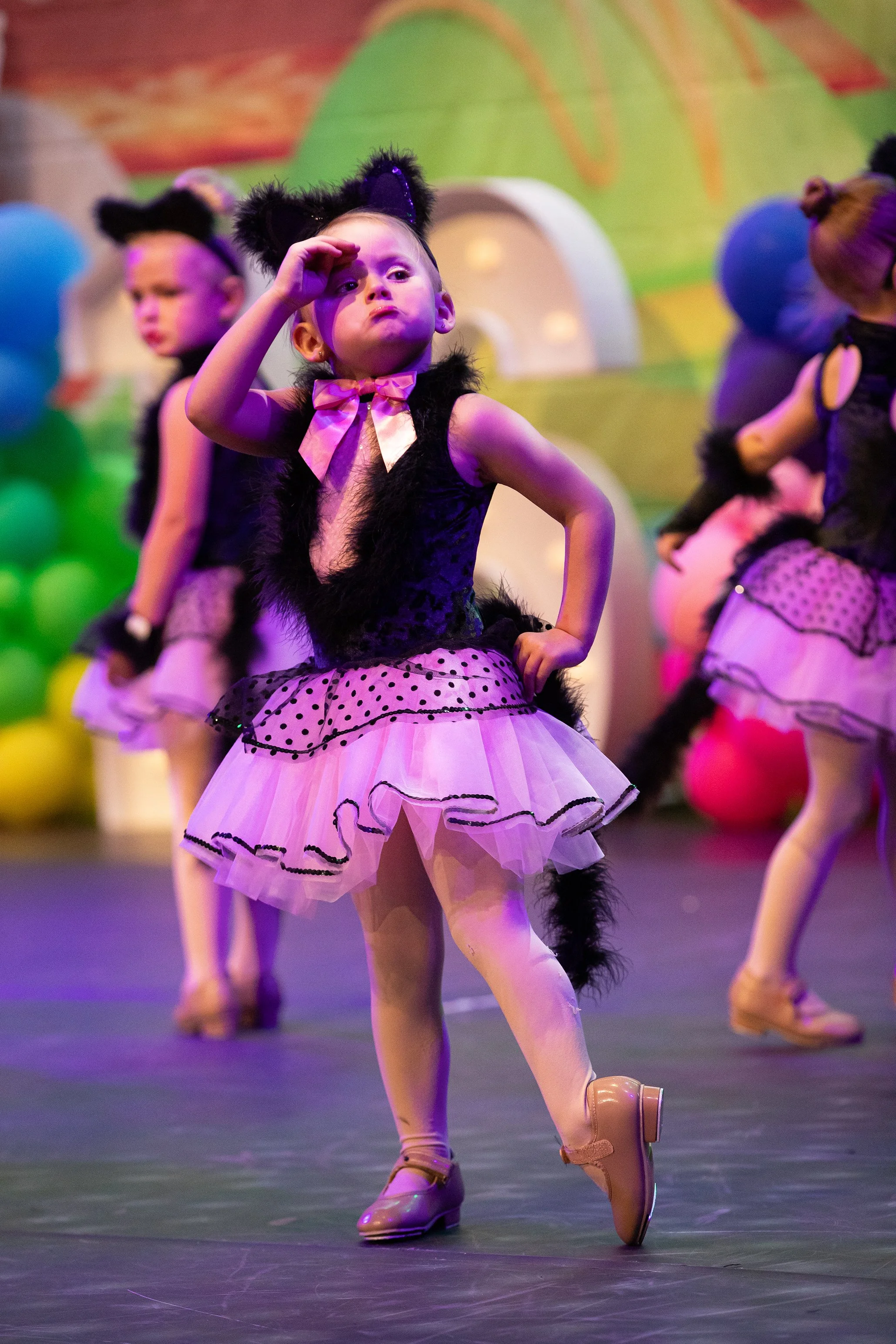 Young girls in black and pink cat costumes performing a dance on stage with colorful balloons and decorations in the background.