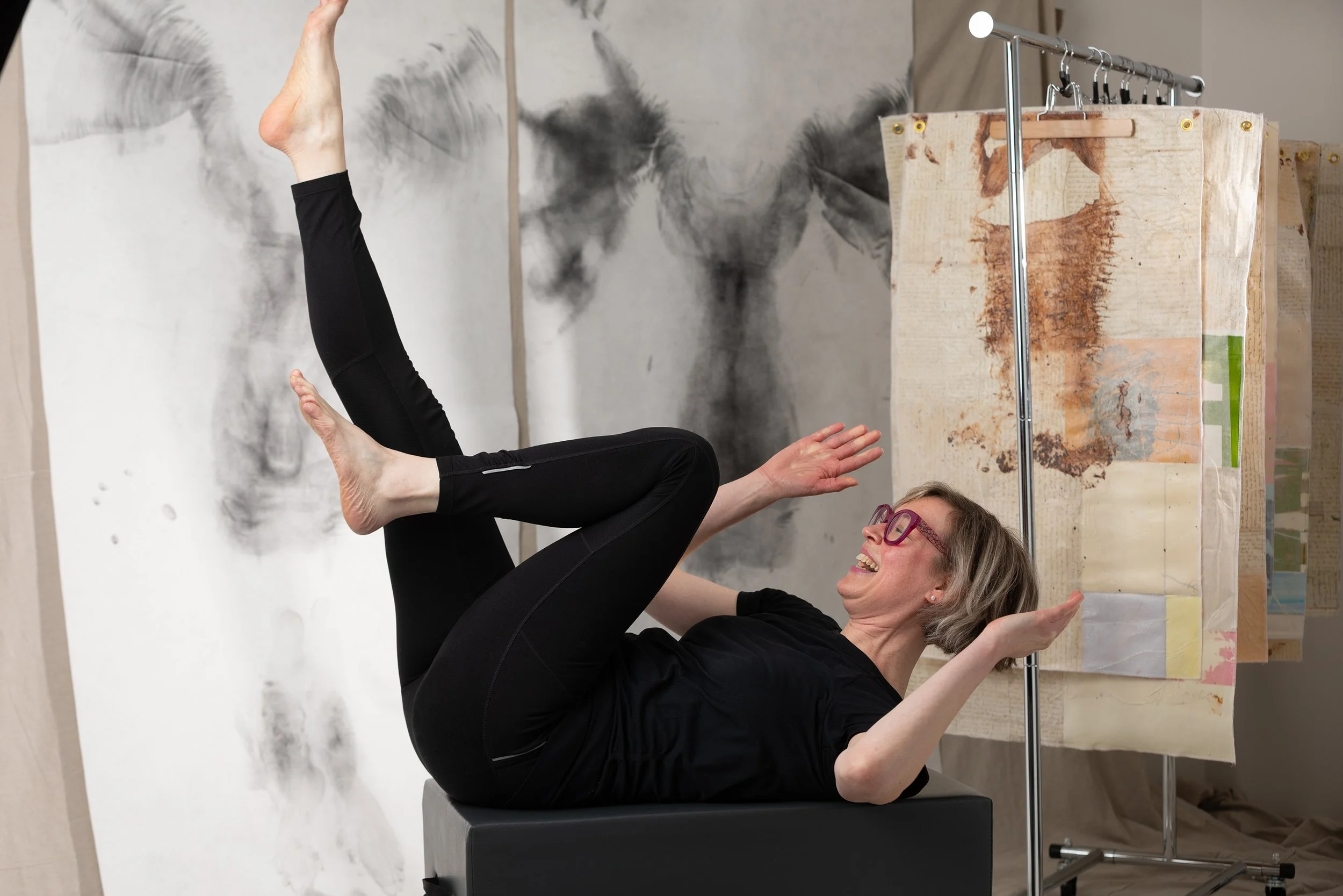 A woman practicing yoga or a stretch in an art studio, with an abstract black and white painting behind her and art supplies nearby.