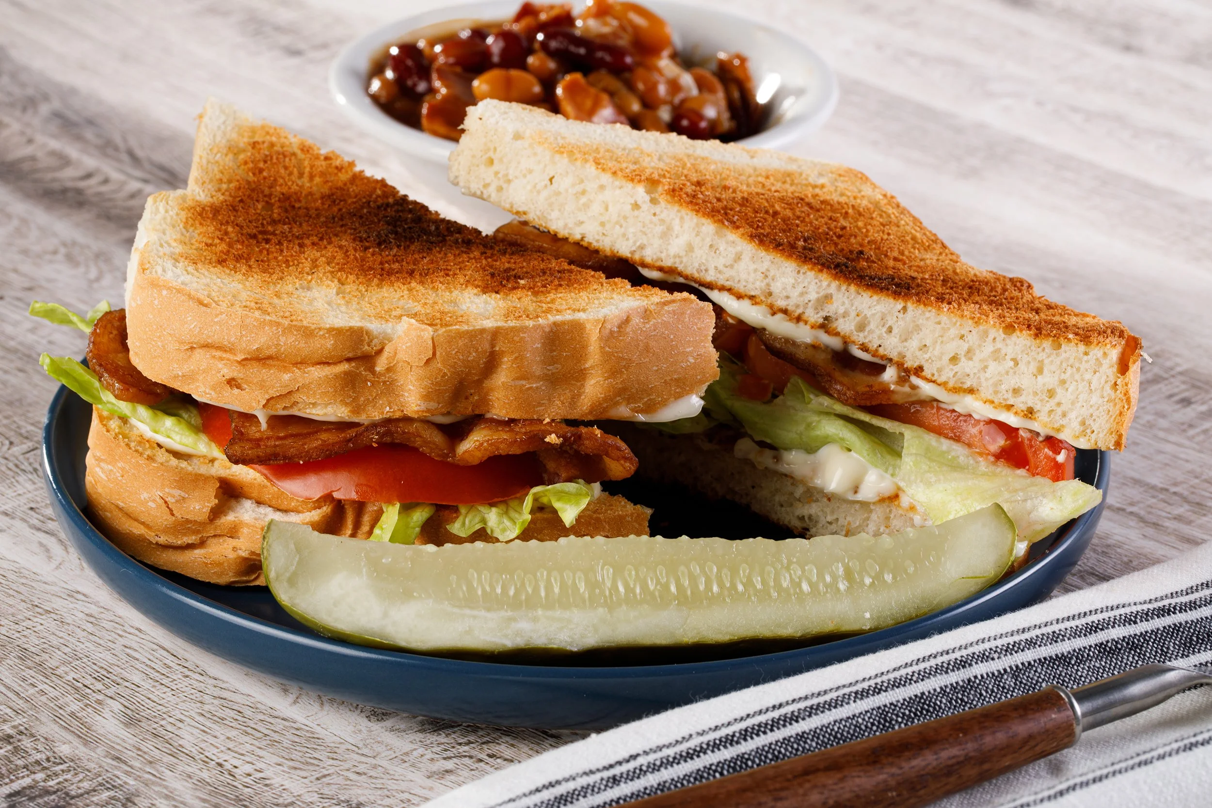 Food photography by Tom Krueger, photographer. Milwaukee, Wisconsin. A sandwich with toasted bread, lettuce, tomato, bacon, and mayonnaise, served with a dill pickle and a side of baked beans in a white bowl on a blue plate.