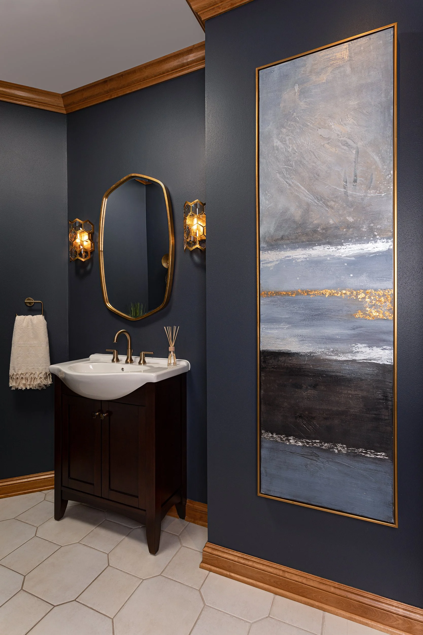 A bathroom with dark blue walls, a white sink with gold fixtures, a large abstract painting with gold trim, hexagonal wall sconces with warm lighting, a mirror with a gold frame, a towel hanging on a gold towel ring, and beige hexagonal floor tiles.