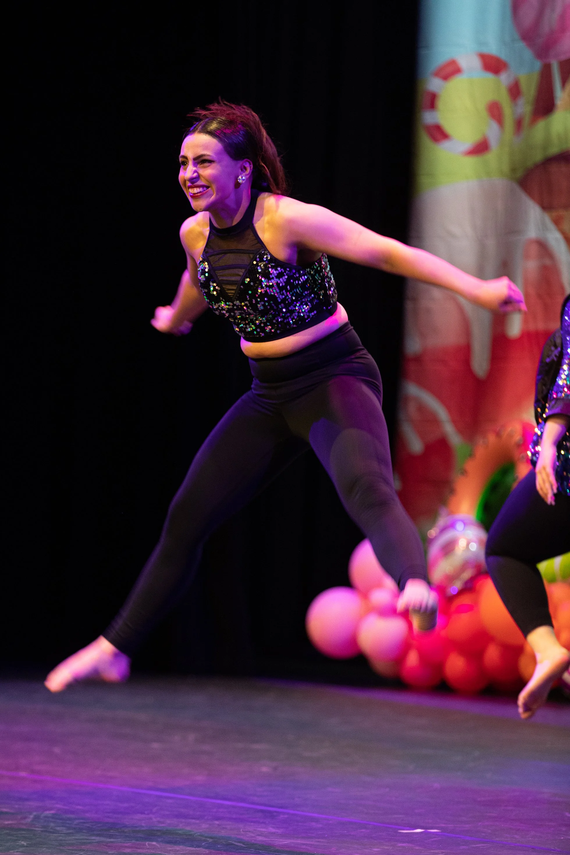 A woman performing a dance move on stage, wearing a black sequined top and black leggings, with a colorful background featuring pink and orange balloons and a cotton candy design.