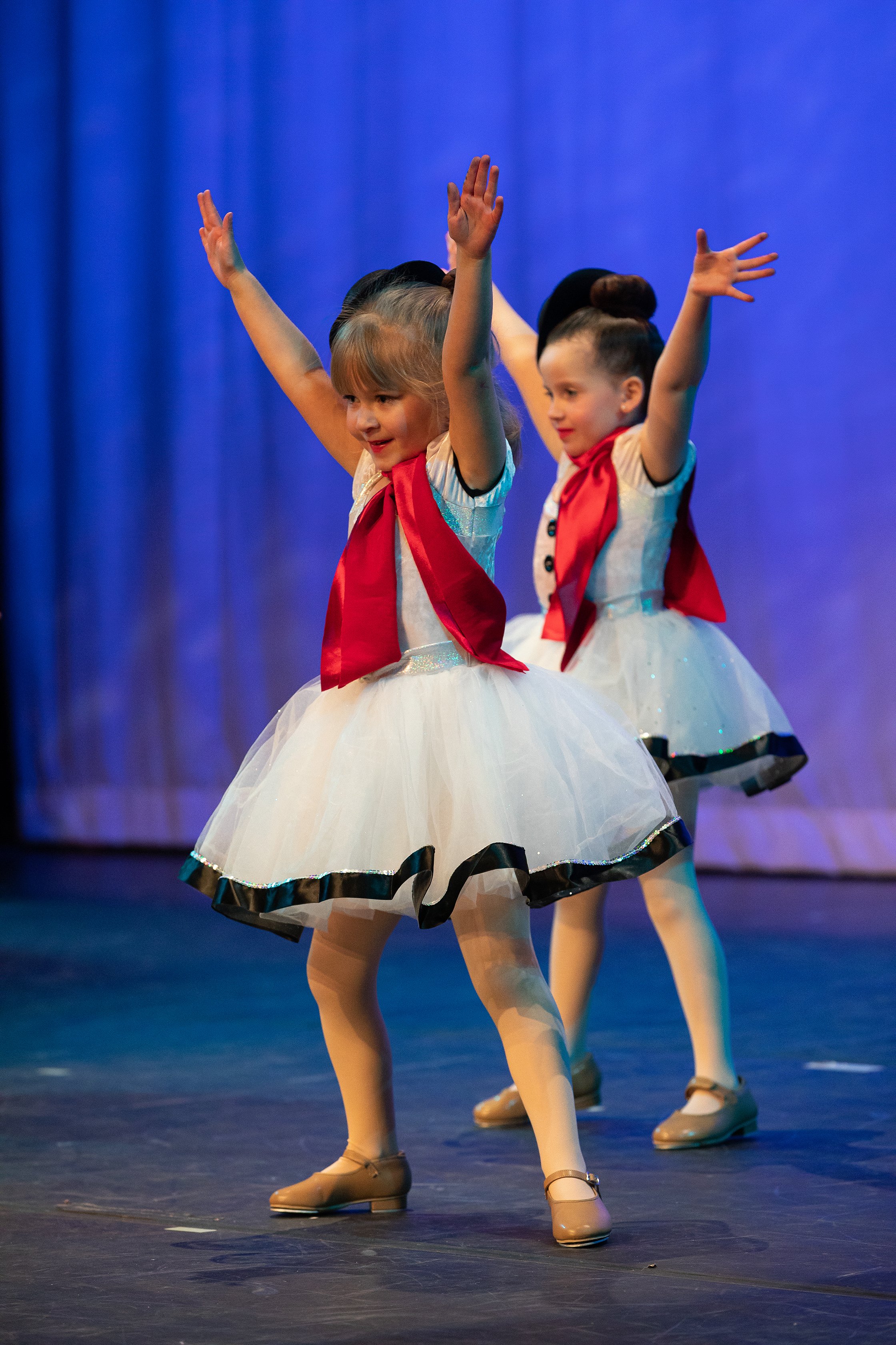 Two young girls in ballet costumes with white tutus, red neckerchiefs, and beige shoes, performing a dance on stage with a blue curtain background.