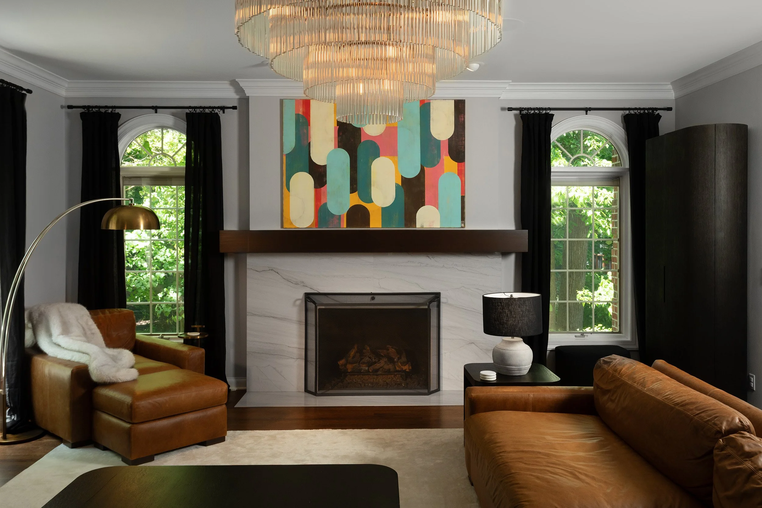 Interior of a living room with a fireplace, large abstract wall art, black curtains, two tan leather sofas, a white lamp, and a chandelier hanging from the ceiling.
