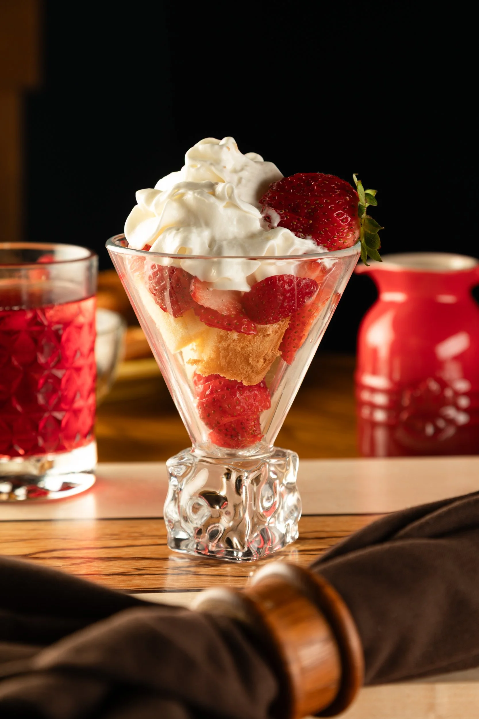 A glass dessert cup filled with strawberries, whipped cream, and cake, garnished with a whole strawberry on top, placed on a wooden surface.