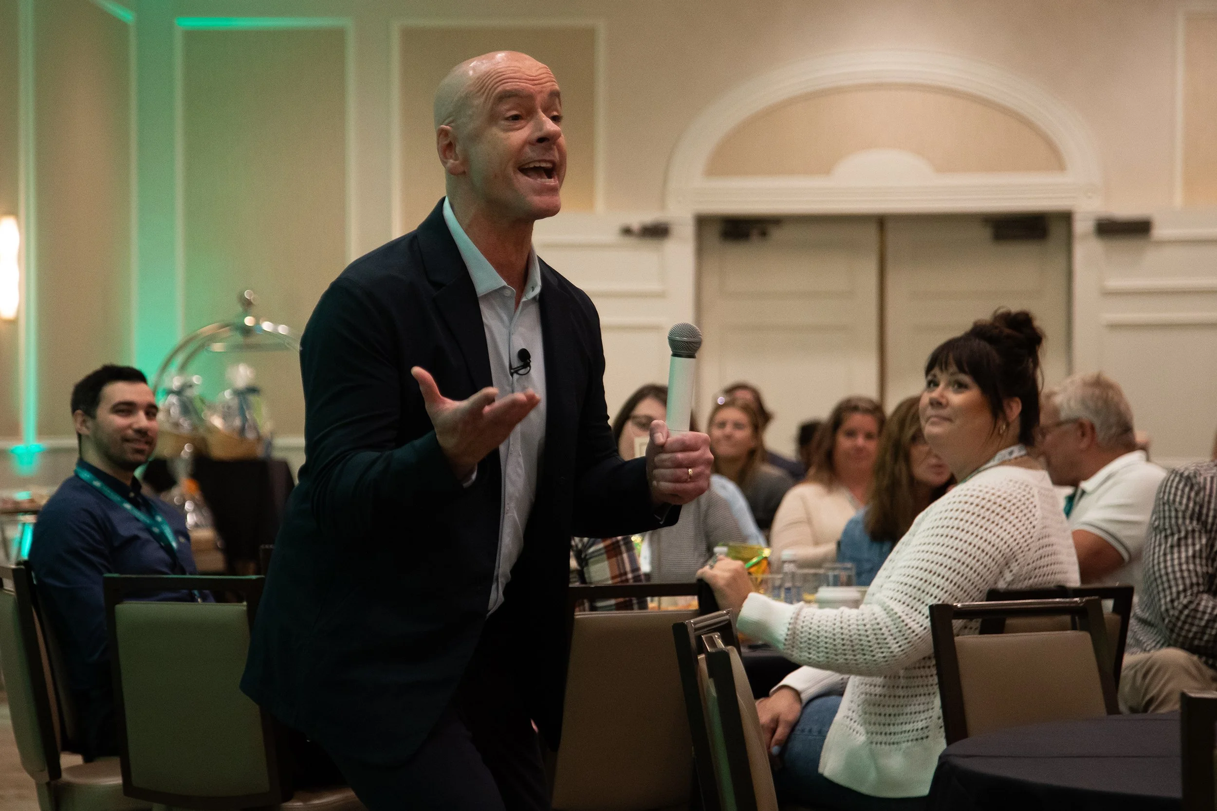A man with a bald head and a suit is speaking into a microphone at an indoor event with many people seated and listening, including a woman in a white sweater.