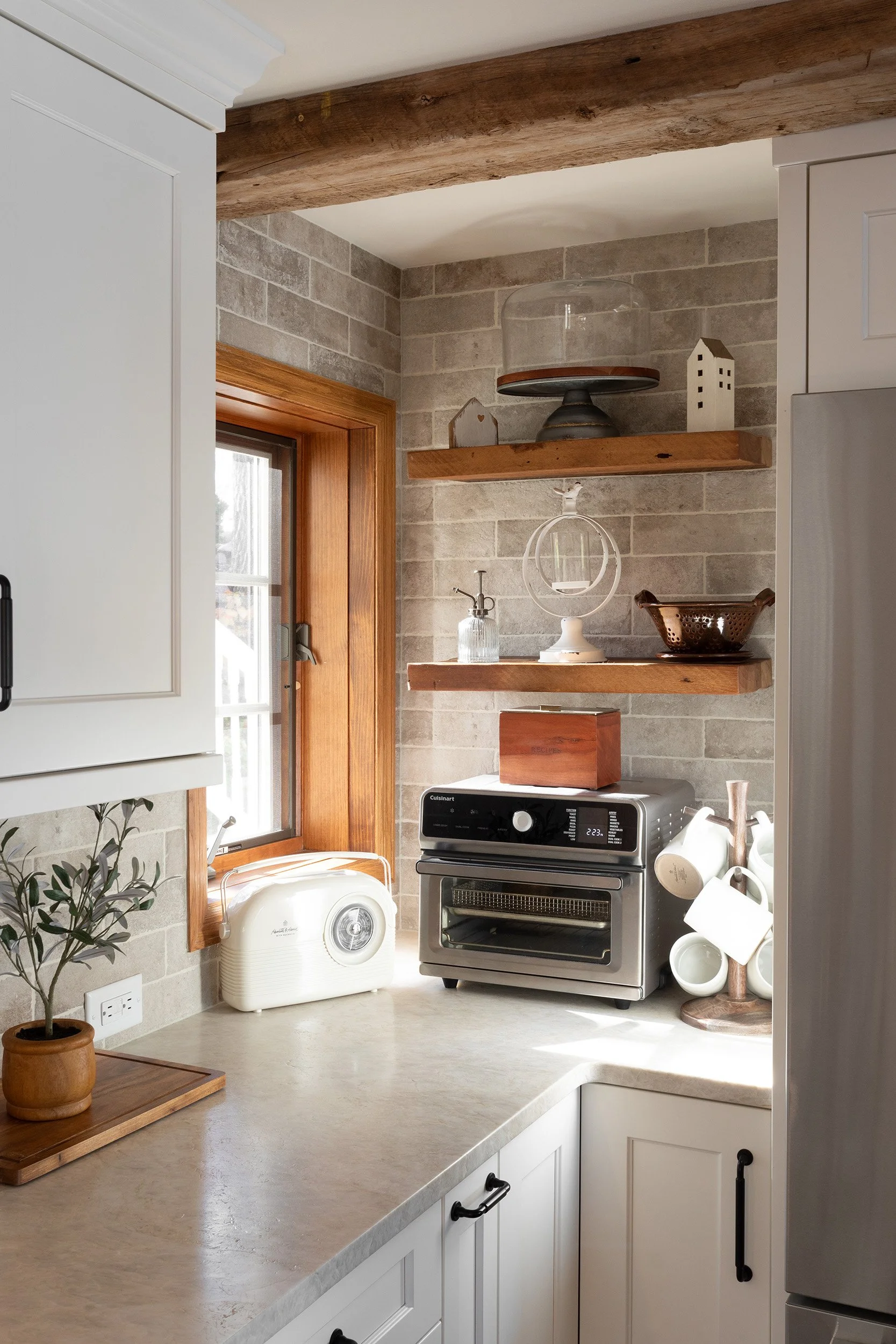 A kitchen with a countertop, white cabinets, a washing machine, brick wall, wooden shelves, and various kitchen items.