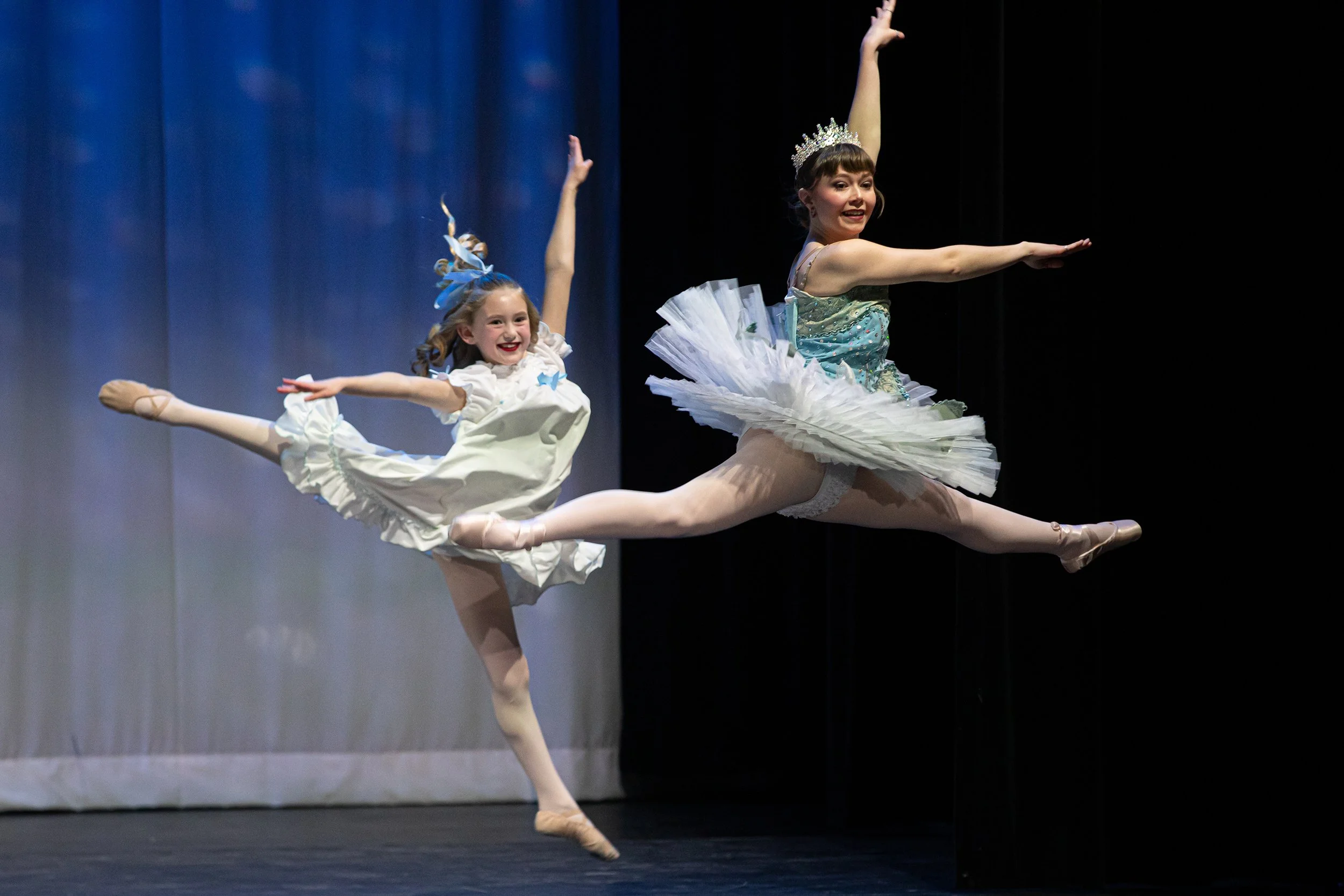 Two young ballet dancers performing a leap on stage, wearing tutus and tiaras, with a blue curtain background.