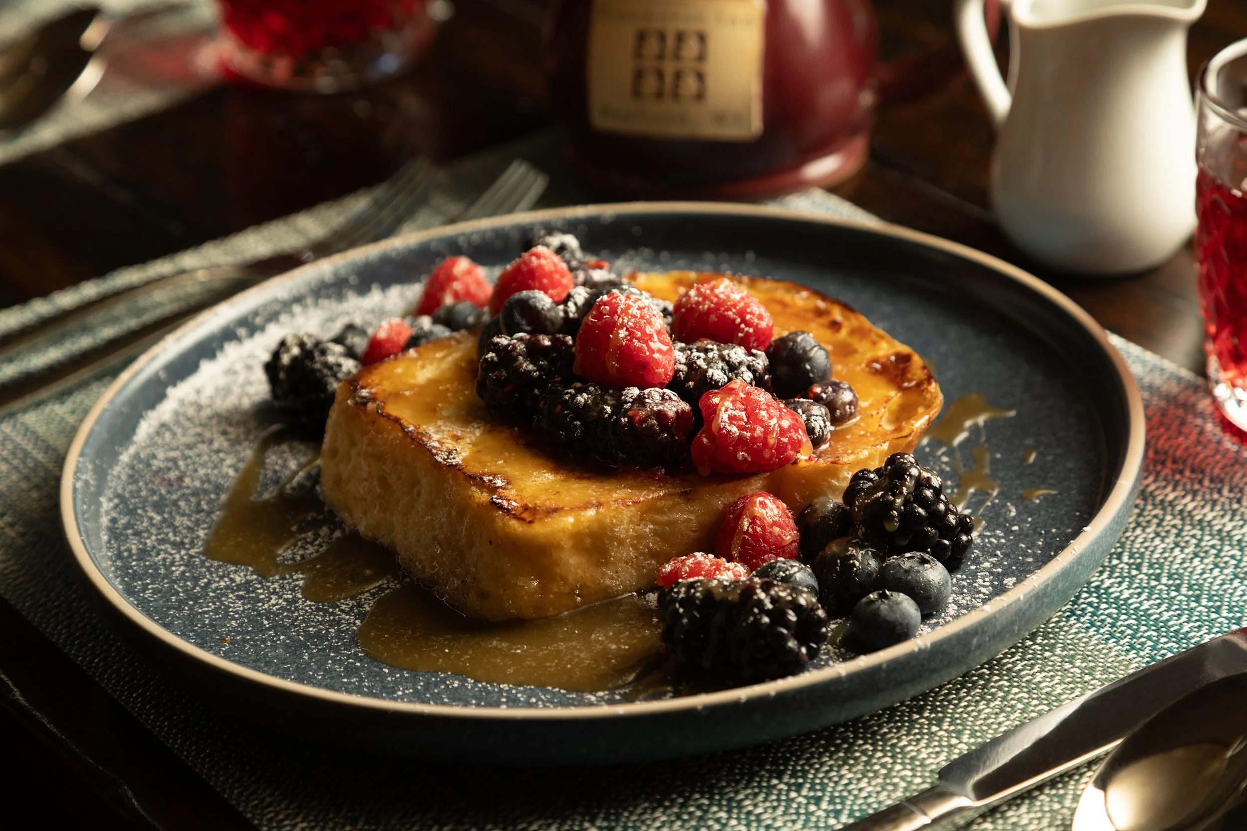 A slice of French toast topped with mixed berries including strawberries, blueberries, and blackberries, dusted with powdered sugar and drizzled with syrup, on a dark ceramic plate.