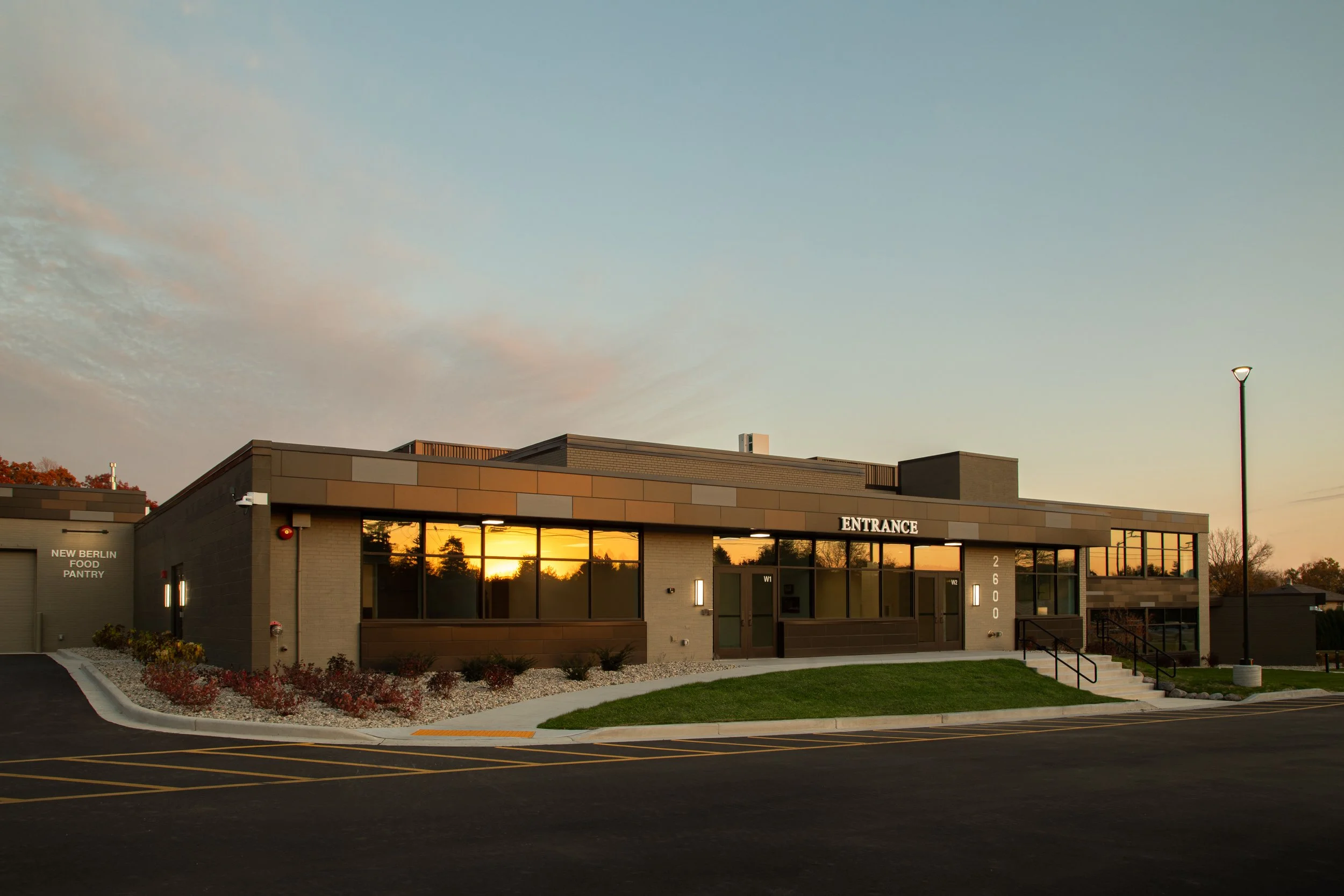 Modern building with large glass windows, illuminated at sunset, labeled 'ENTRANCE' with street number 2600, well-maintained landscaping, parking lot in foreground, and a lamppost to the right.