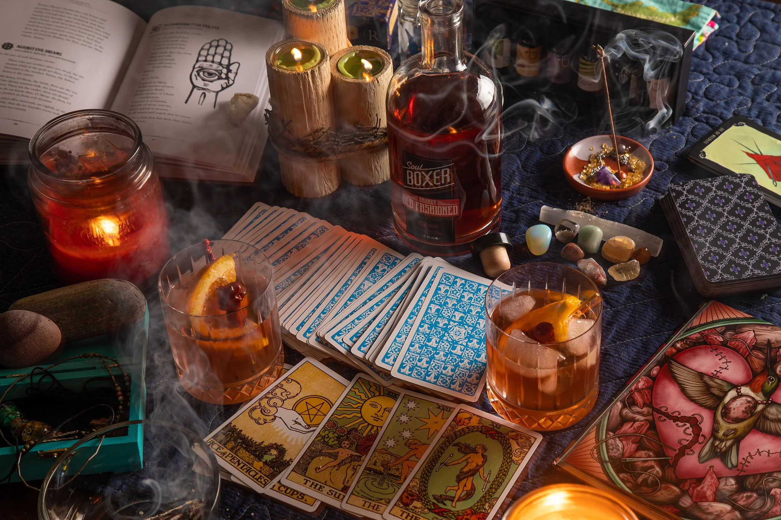 A table set with tarot cards, candles, a bottle of liquor, glasses of iced drinks, crystals, jewelry, and various mystical items for a ritual or spiritual practice.