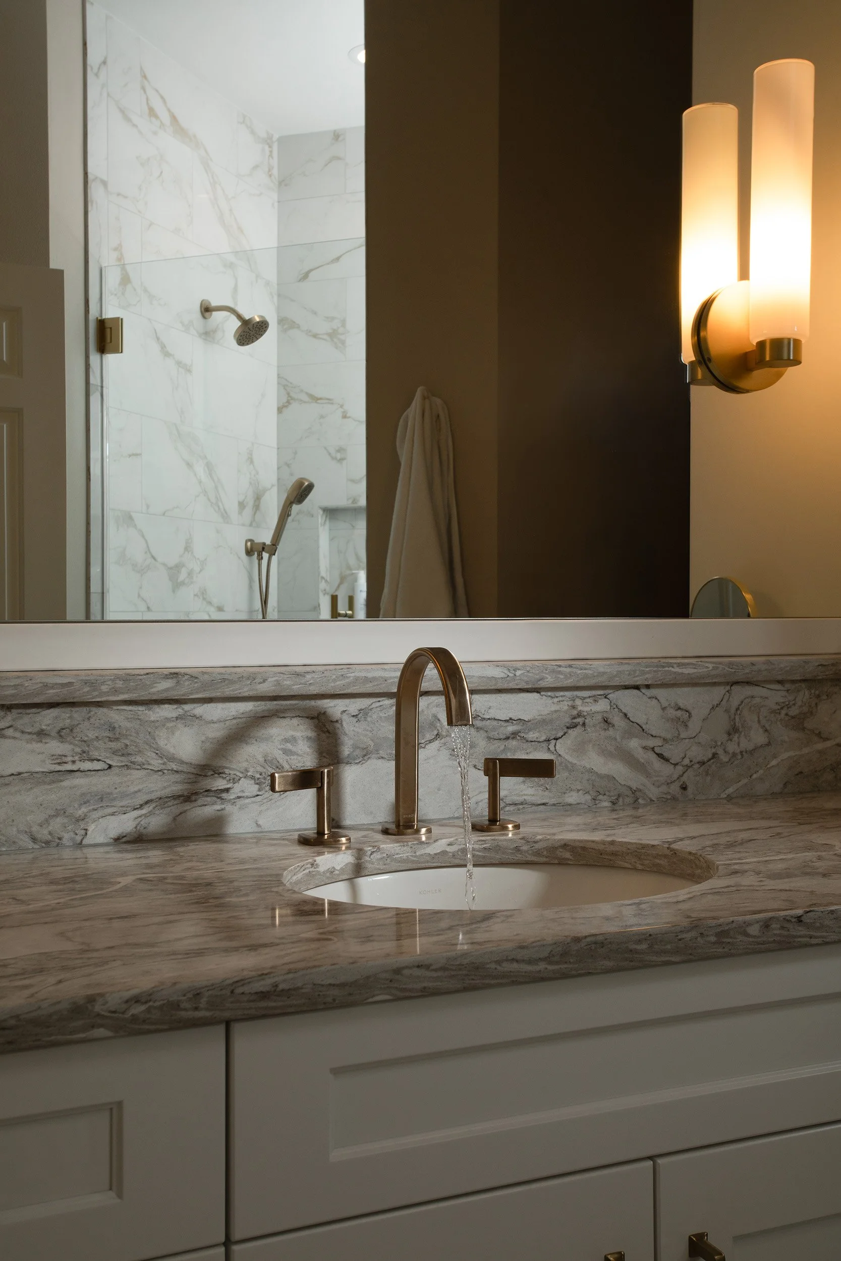 Close-up of a bathroom mirror, marble countertop with a sink and gold-tone faucets, and a wall-mounted light fixture with two vertical frosted glass shades; in the background, a shower with marble tiles, a handheld showerhead, and a wall hook with a 