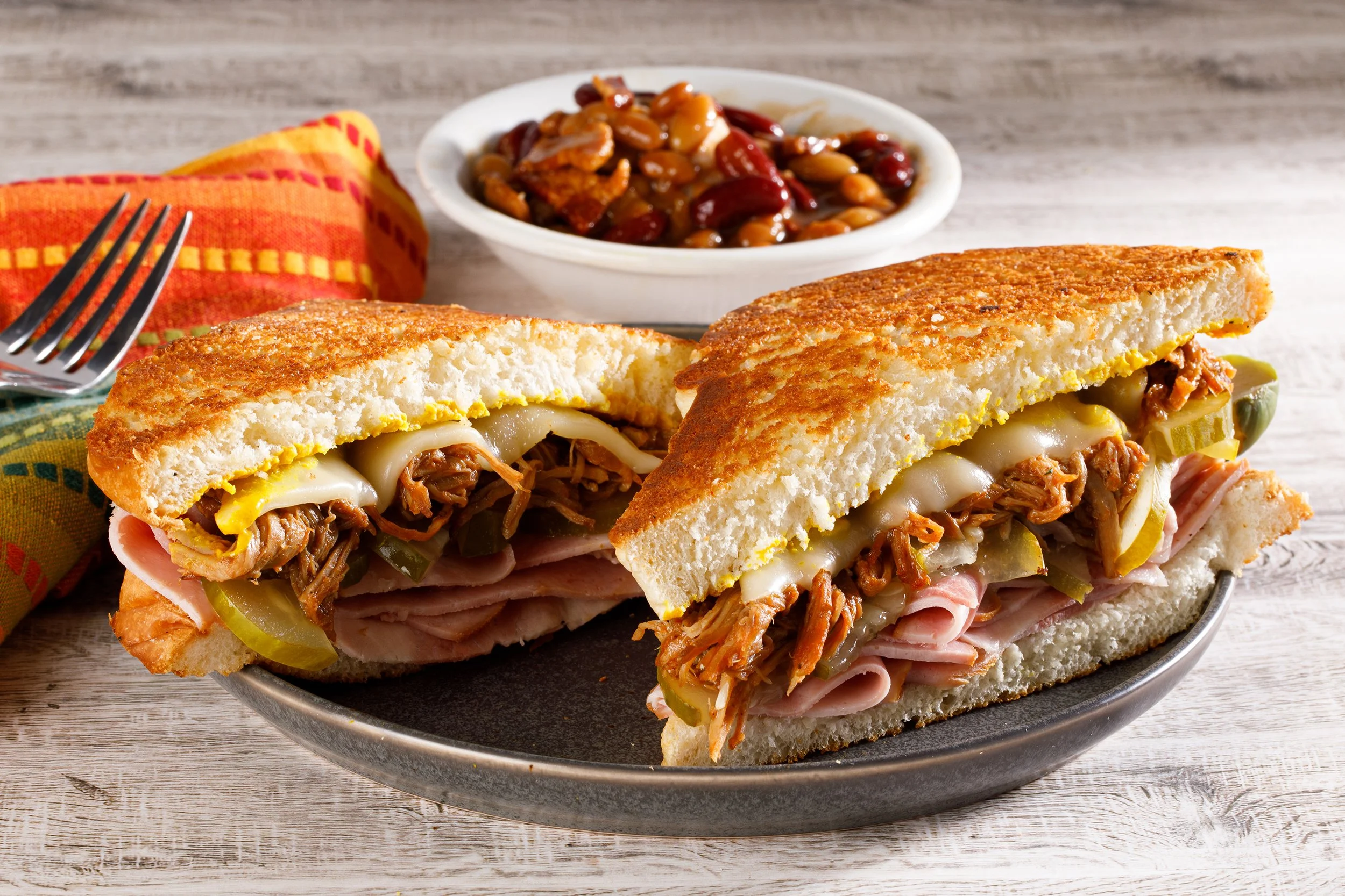 Food photography by Tom Krueger, photographer. Milwaukee, Wisconsin. A plate with a sandwich filled with roasted meat, pickles, and cheese, alongside a small bowl of baked beans, on a wooden surface with a colorful napkin.