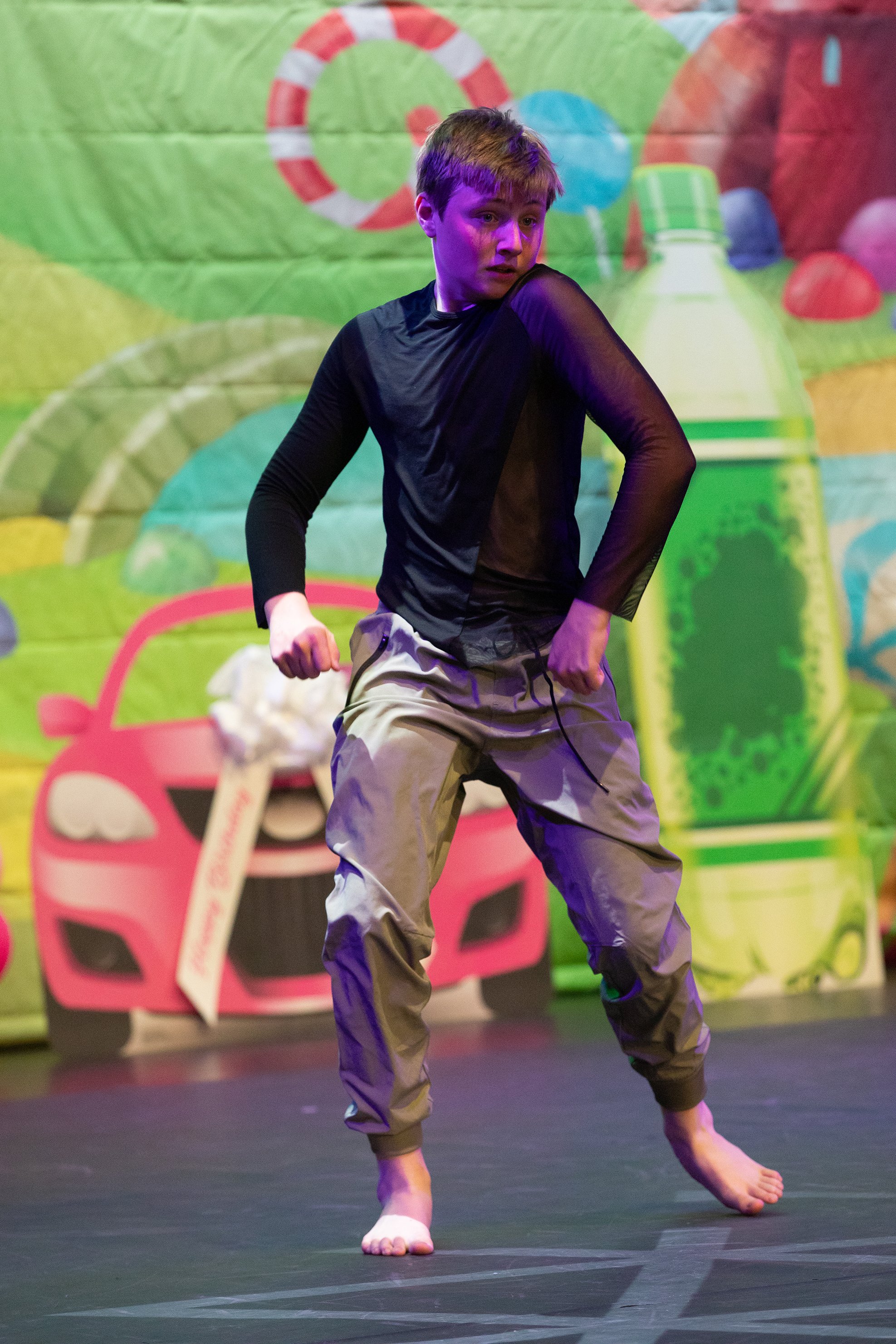 A young man performing a dance on a black stage, with a colorful, playful backdrop featuring cartoonish illustrations of toys and a pink toy car.