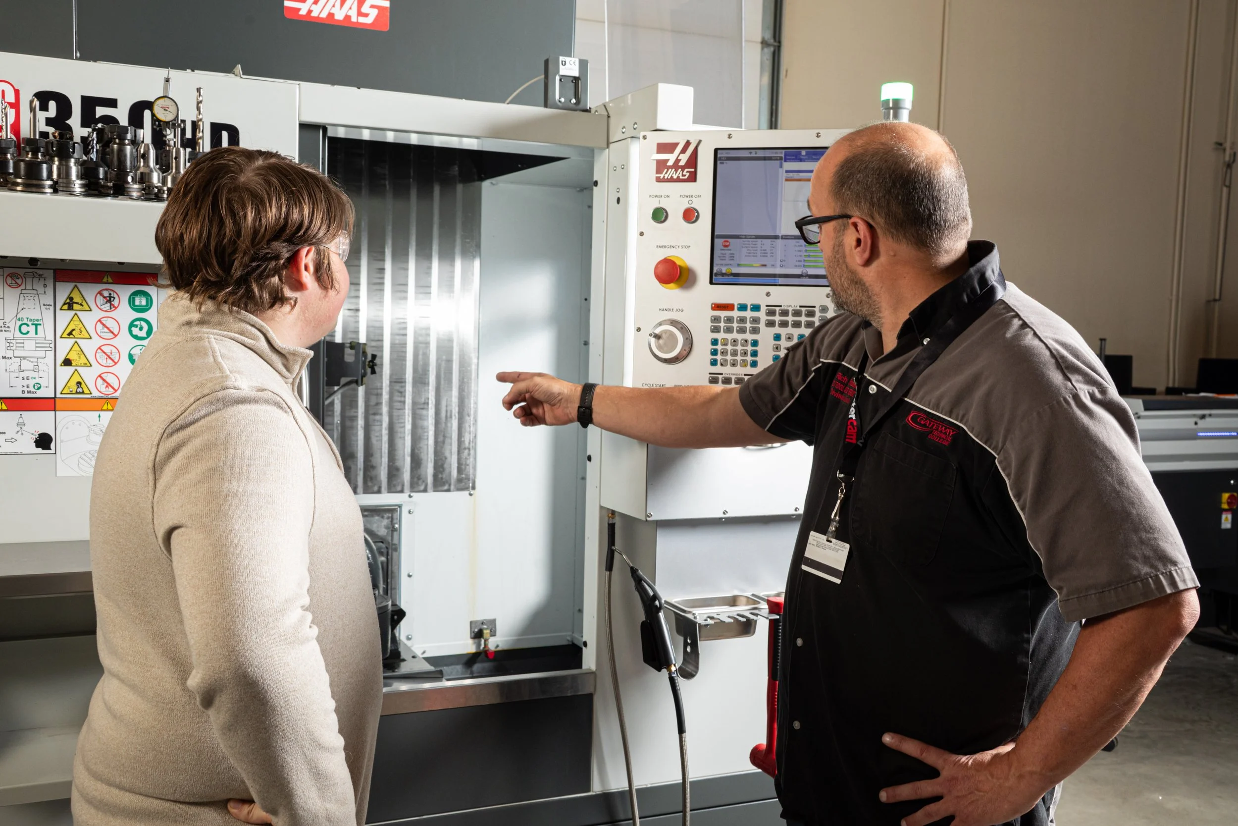Two men, one with glasses, and one with a cap, are inspecting industrial machinery with a computer control panel in a factory setting.