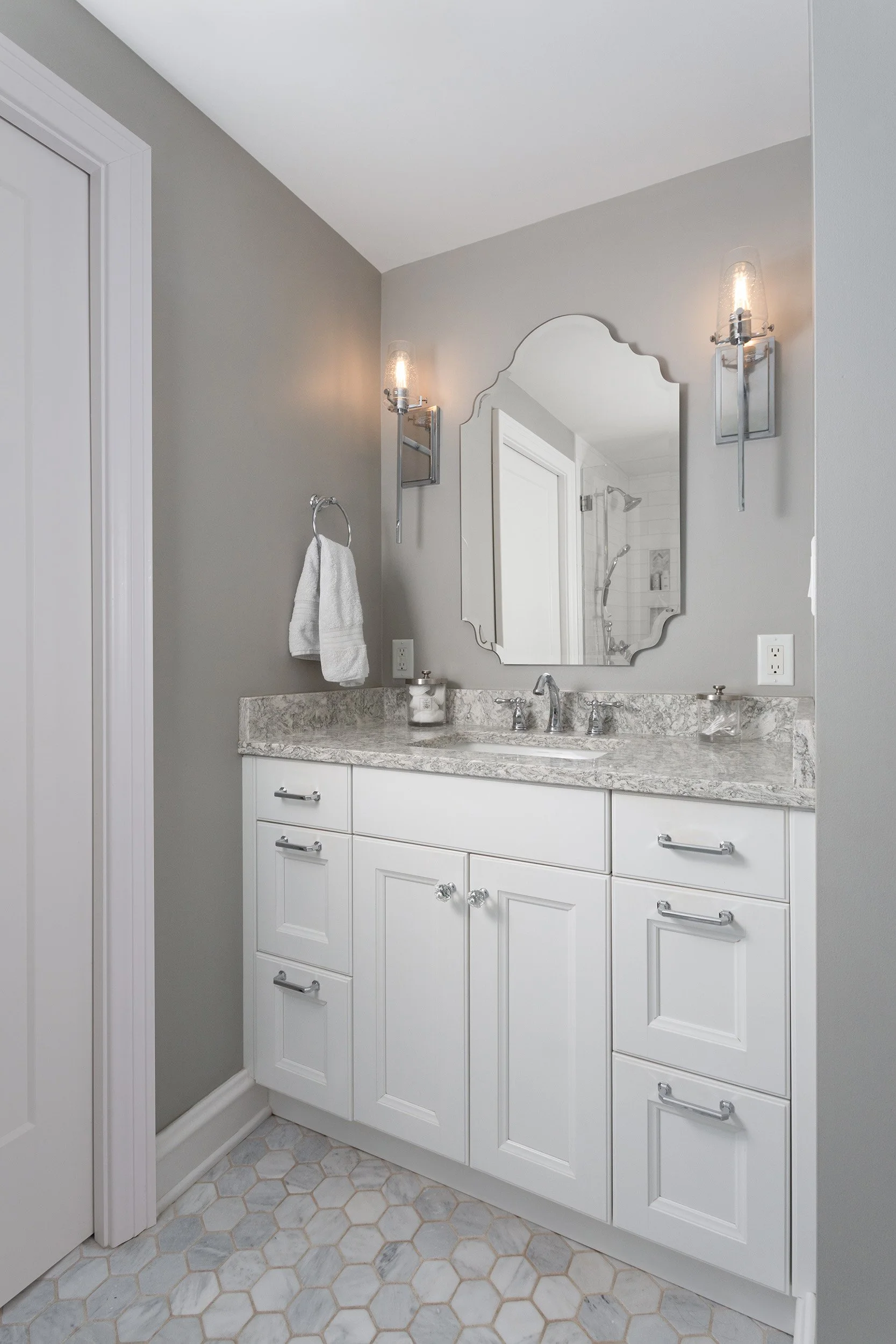 A bathroom vanity with a white cabinet, marble countertop, and silver hardware. There are two mirrors, one being decorative and irregularly shaped. Wall-mounted light fixtures are on either side of the mirror. The reflection shows a shower with a gla