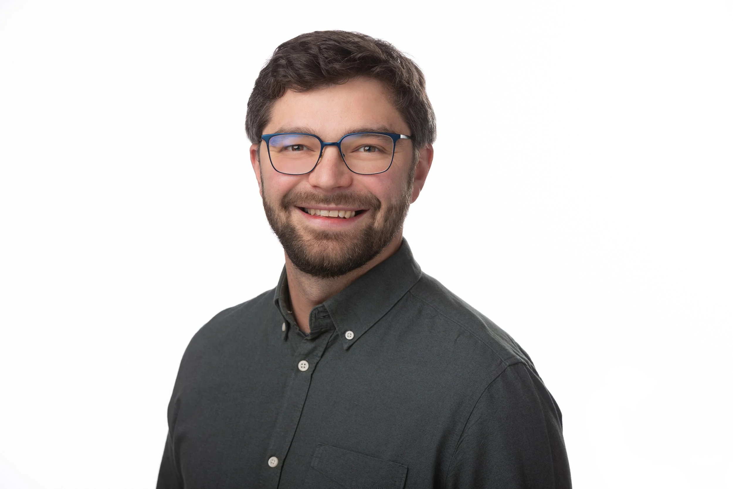 Portrait of a smiling man with glasses, dark hair, and a beard, wearing a dark button-up shirt, against a white background.