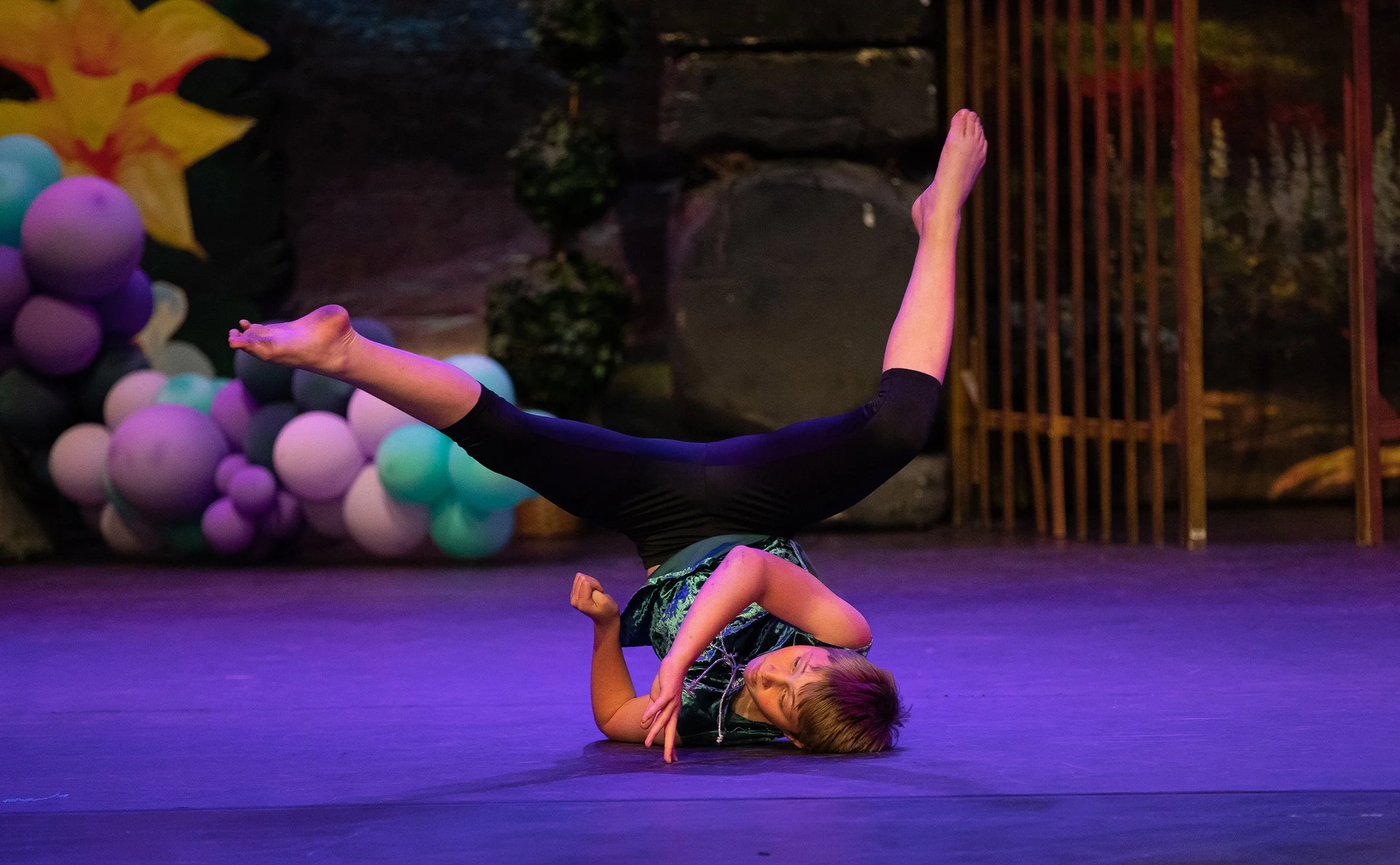 Young girl performing a dance move on stage, with balloons and rocks in background.