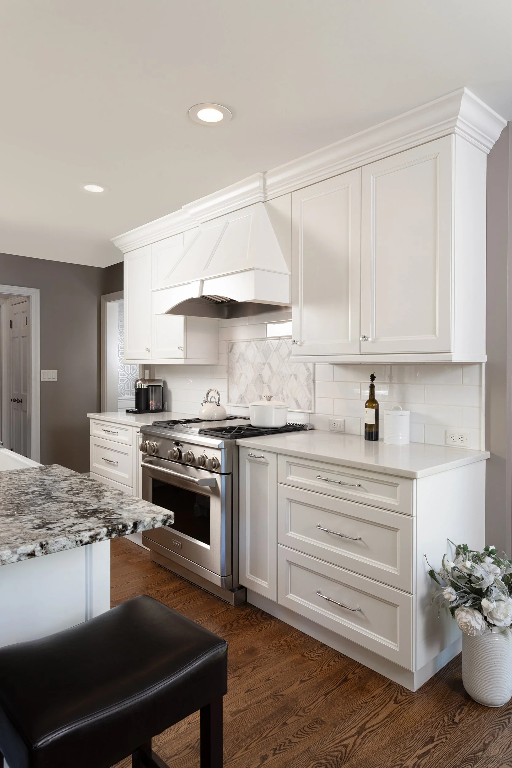 A modern kitchen featuring white cabinetry, a stainless steel stove, a granite countertop island, and hardwood floors. There is a white teapot on the stove, a wine bottle with a glass, and flowers in a white vase on the counter. Recessed lighting and
