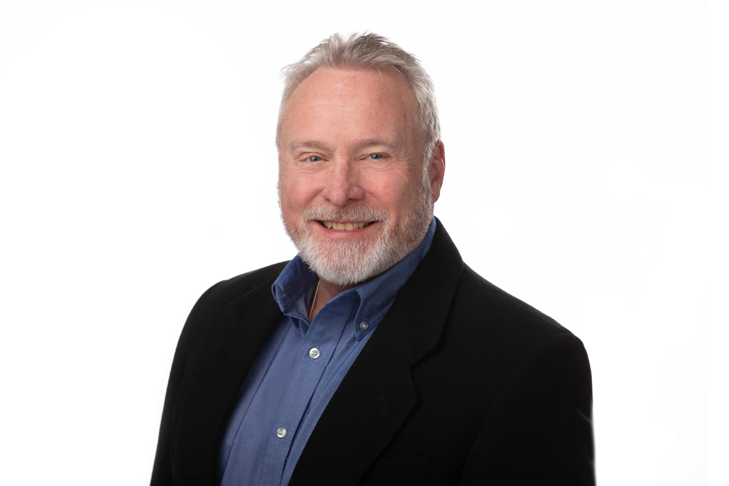Headshot of a smiling middle-aged man with gray hair and beard wearing a navy blue shirt and black blazer against a white background