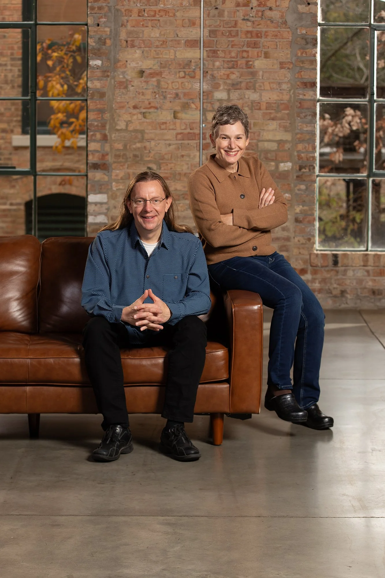 A man and woman posing indoors in front of a brick wall with large windows, the man sitting on a brown leather couch and the woman sitting on its armrest. Both are smiling, with the woman crossing her arms.