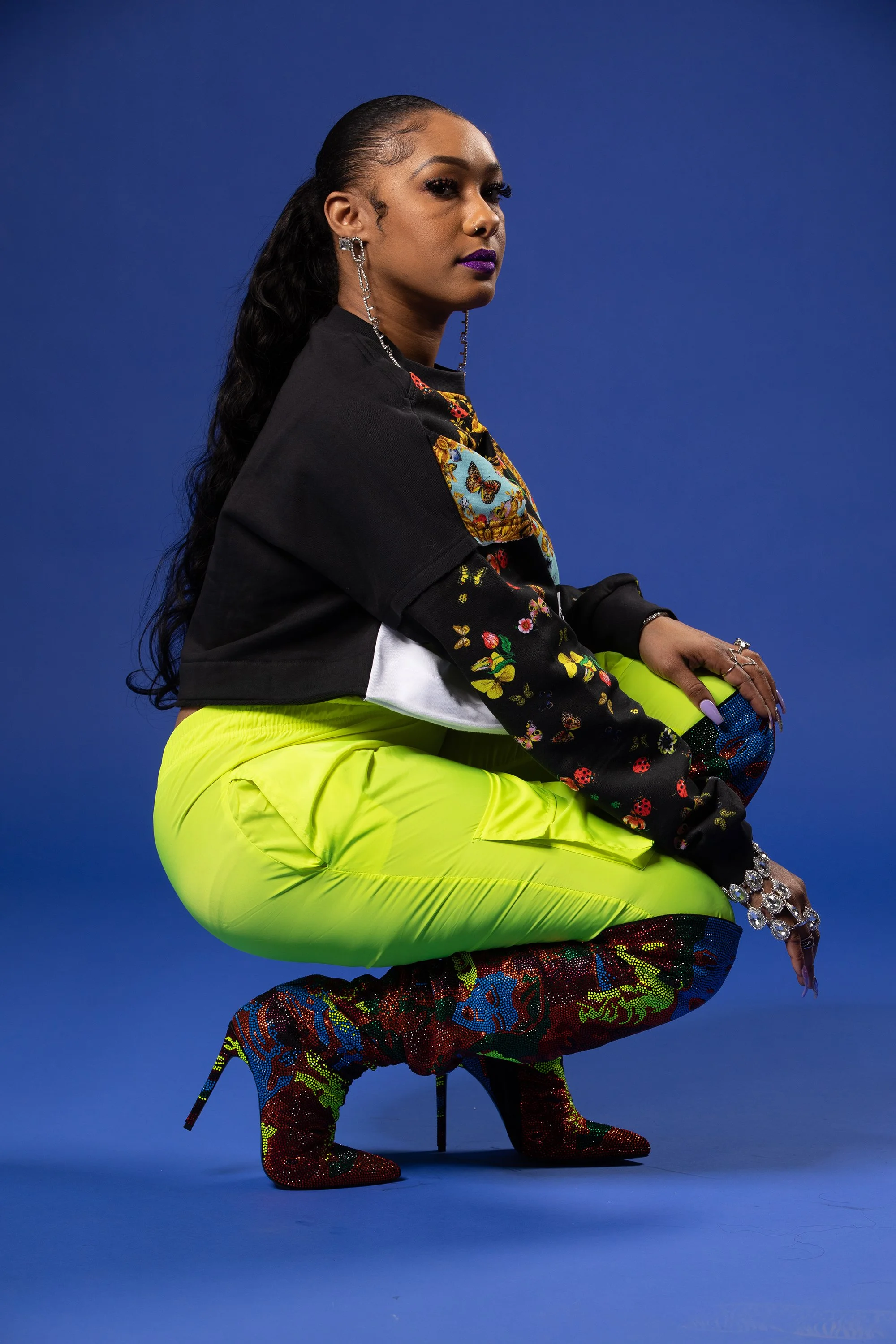 Woman with long wavy hair and purple lipstick, wearing large dangling earrings and a black shirt with colorful floral and butterfly designs, neon yellow pants, patterned thigh-high boots, and jewelry, posed against a solid blue background.