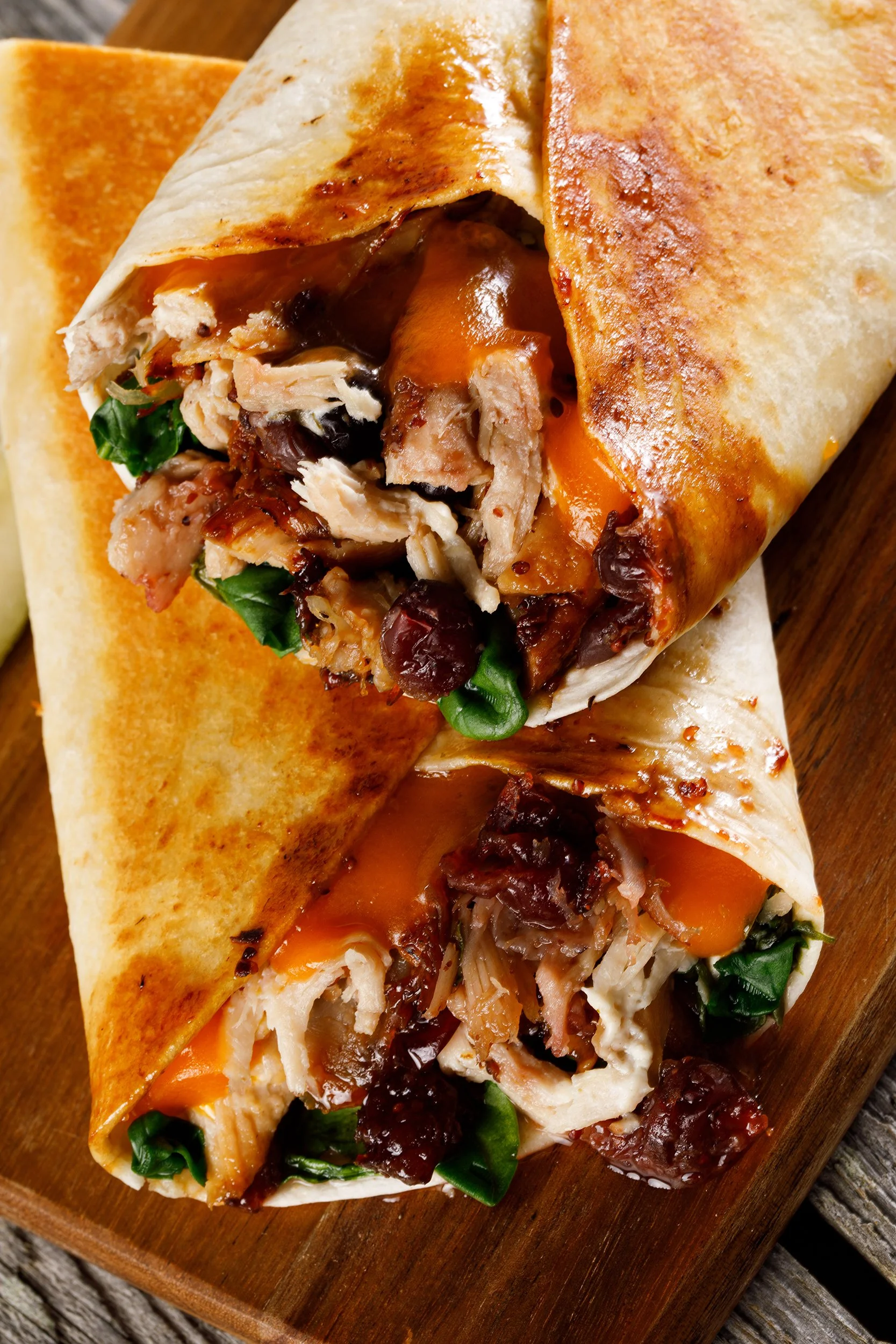 Close-up of a burrito with a toasted flour tortilla filled with shredded chicken, spinach, black beans, cheese, and pulled pork, served on a wooden board.