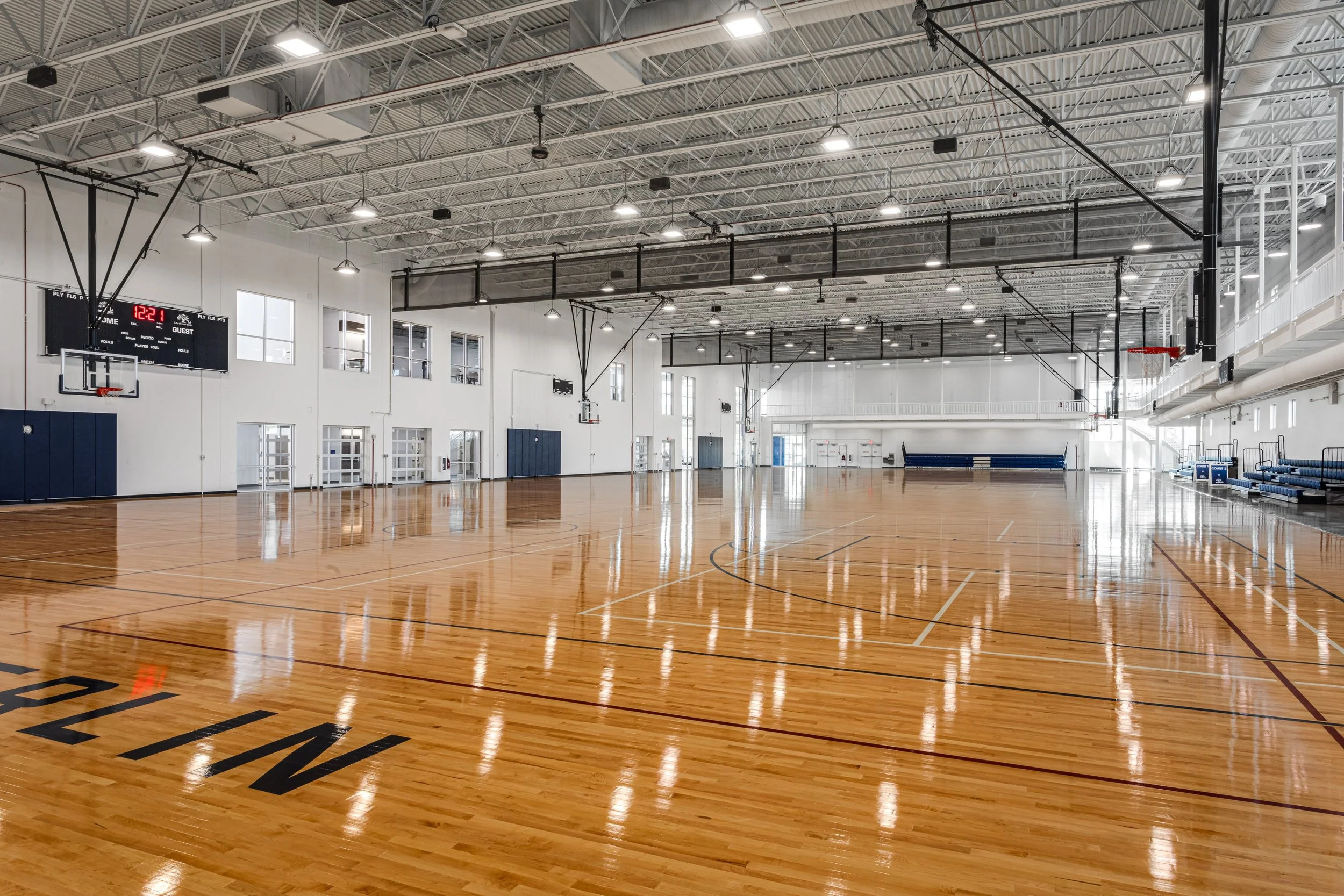 Empty indoor basketball gym with polished wooden floor, multiple basketball hoops, large windows, and seating area on the right.
