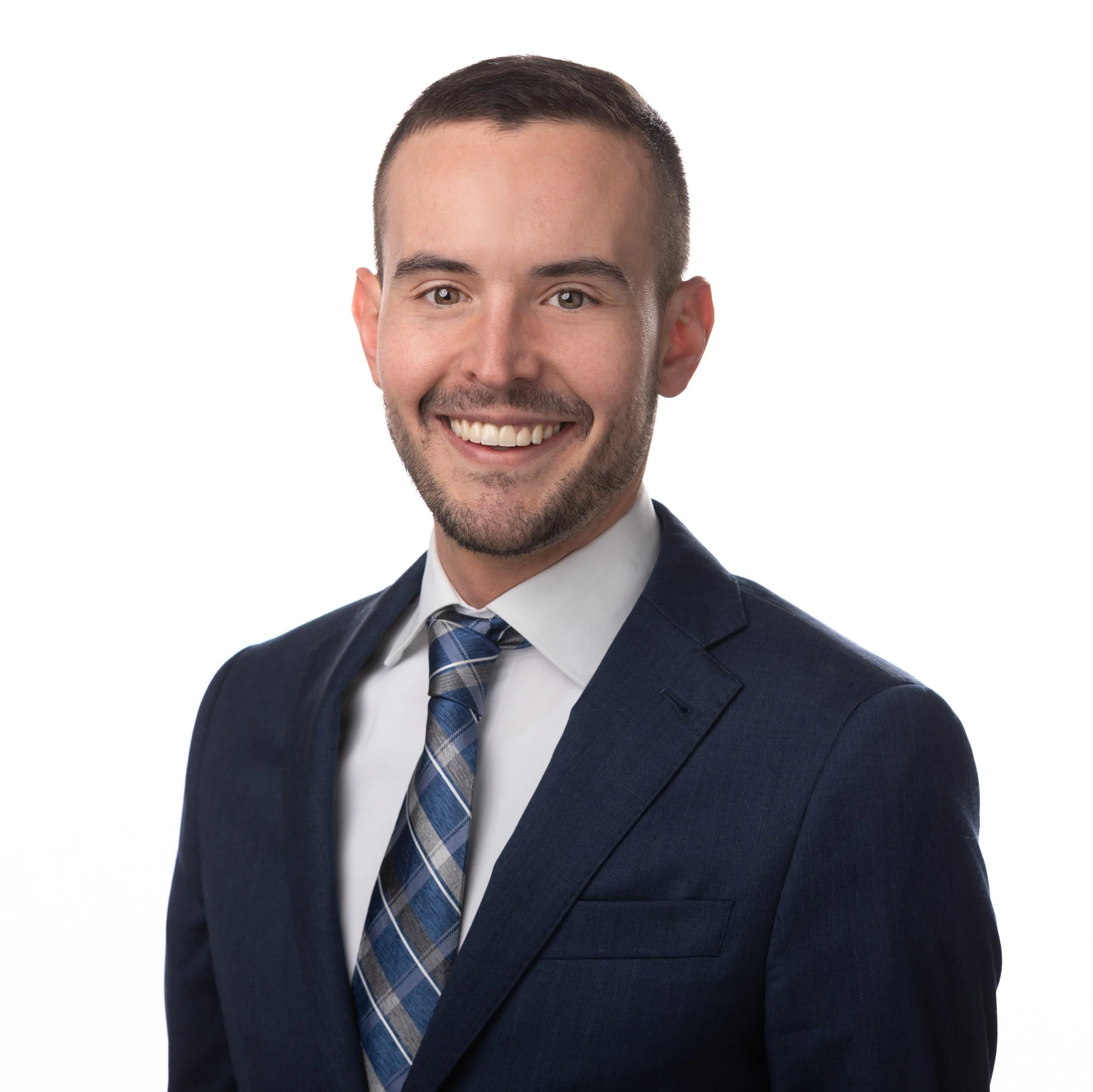 Close-up of a smiling man with a neatly trimmed beard, wearing a navy suit, white shirt, and blue plaid tie, against a plain white background.