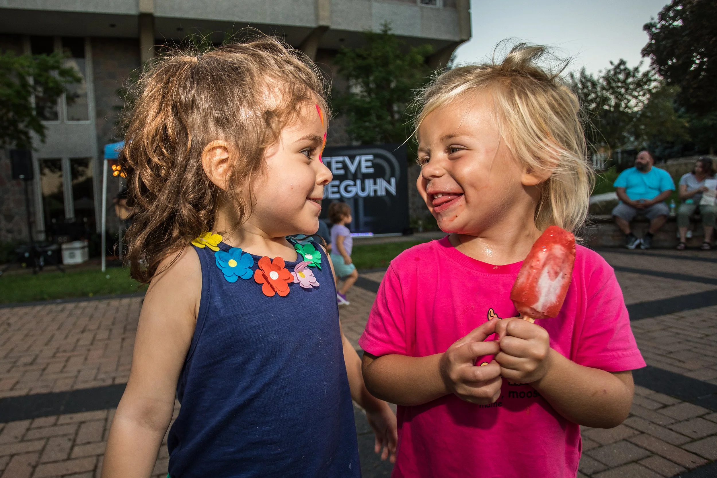 Two young girls interact closely outdoors, one holding a strawberry-flavored ice pop, with a background of people seated and a sign that reads 'Steve Seguhn' at dusk.