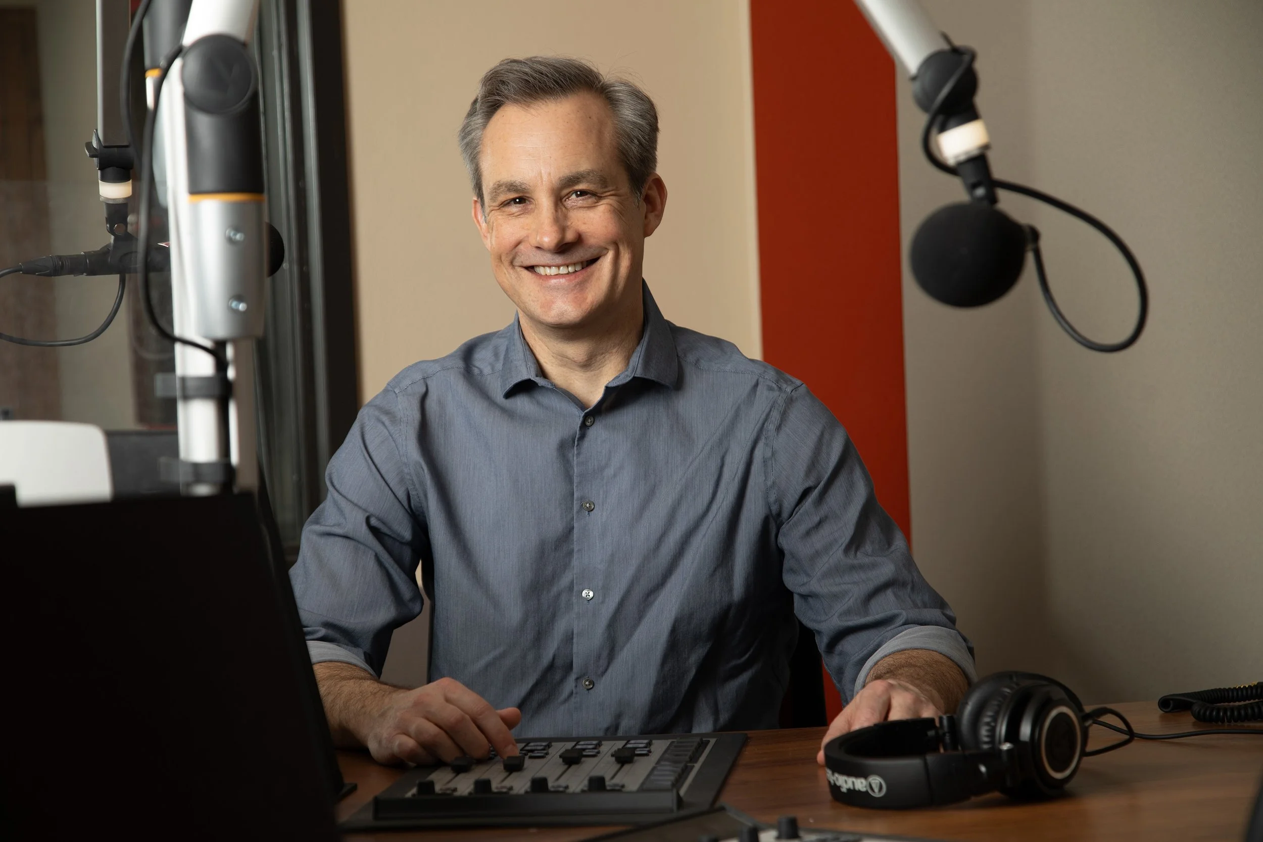 A smiling man with gray hair, wearing a gray button-up shirt, sitting at a desk with audio equipment including headphones, a microphone, a computer, and a recording device in a studio or office setting.