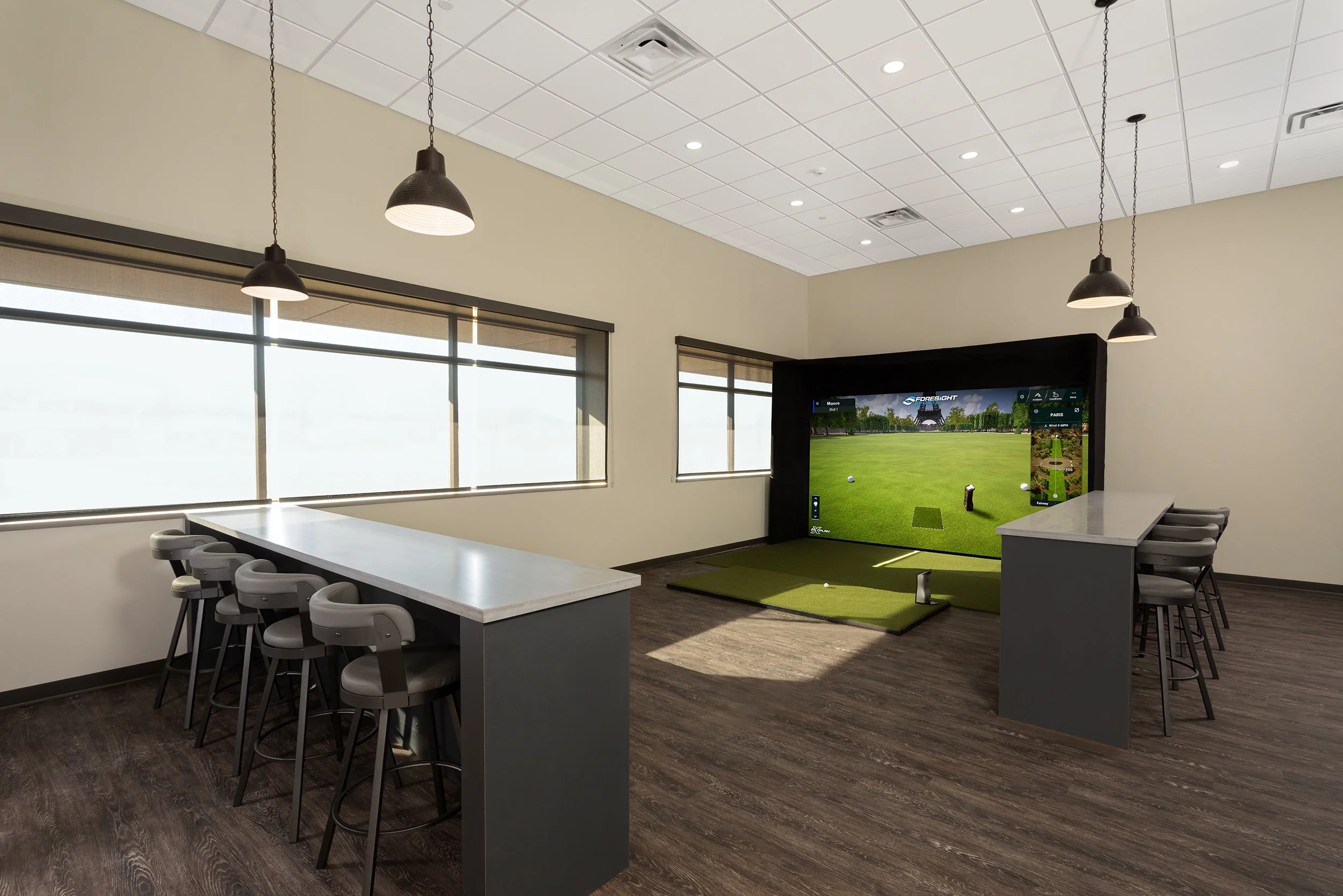 Indoor golf simulation room with a putting green, a large screen displaying a golf course, bar-height chairs around a counter, and ceiling pendant lights.
