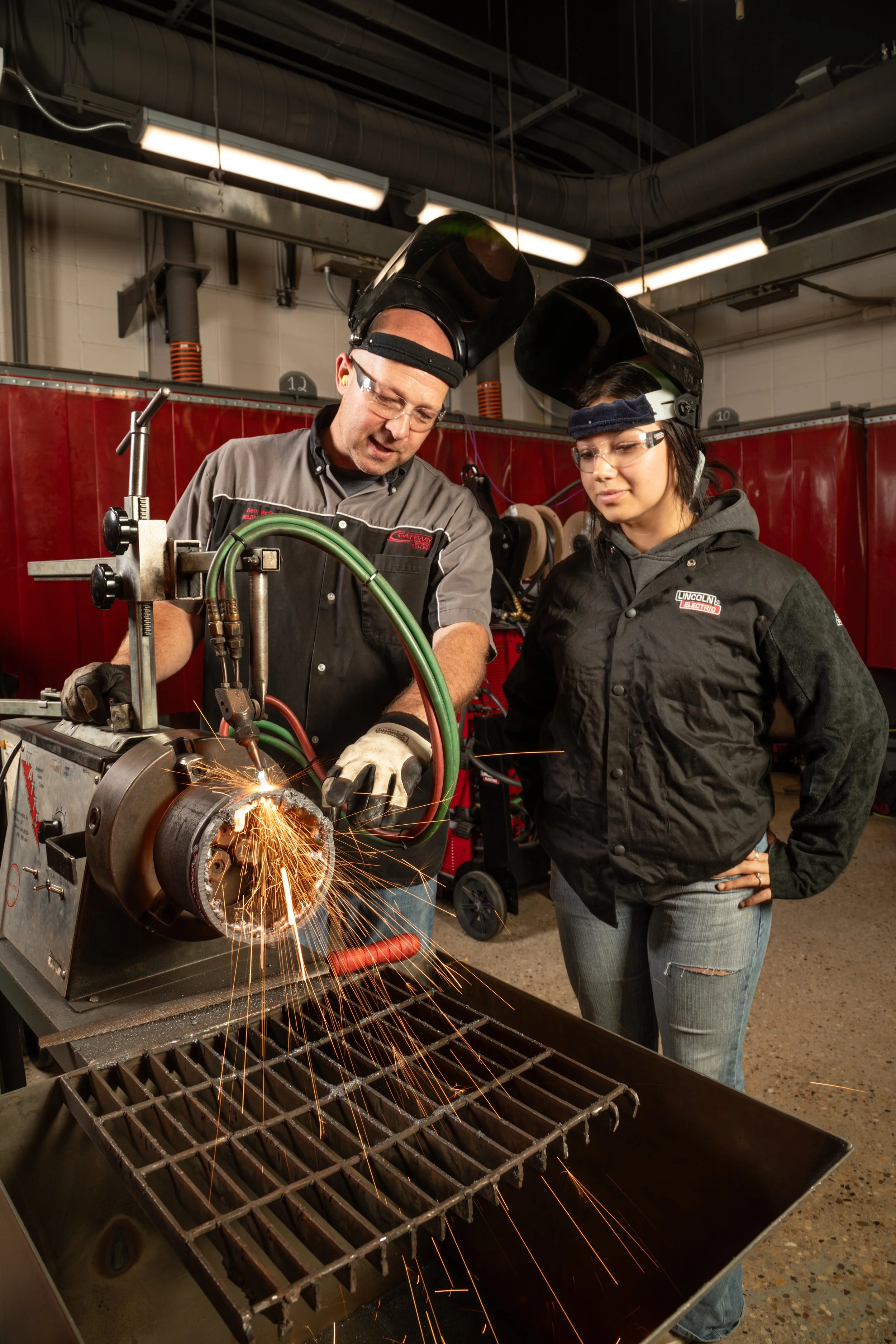 Two people wearing protective gear, including welding helmets and safety glasses, working together on a welding project in a workshop. Sparks are visible as the welding torch is used on a metal pipe.