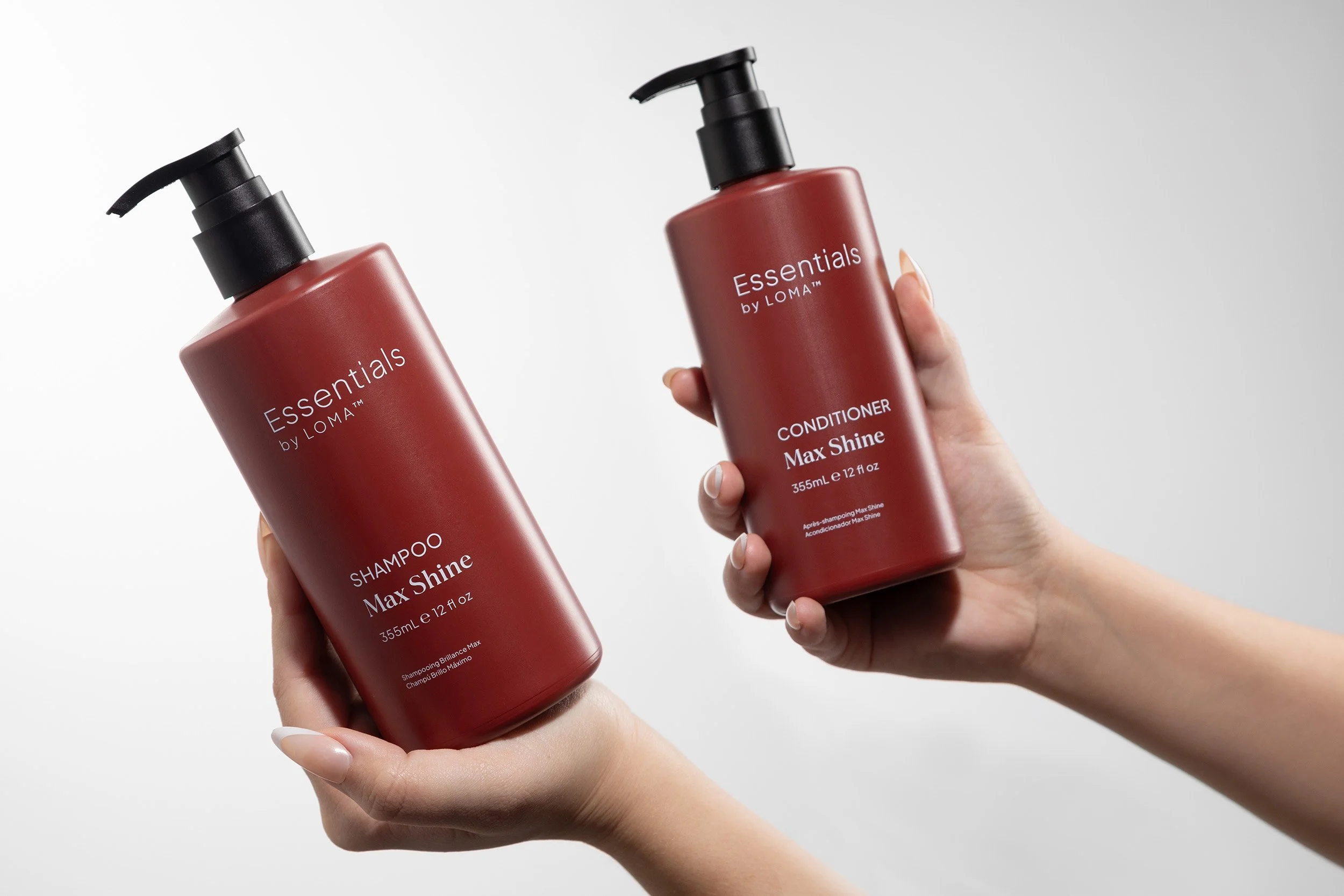 Two hands holding red bottles of hair care products, shampoo and conditioner, labeled 'Essentials by LOMA Max Shine'. The background is light gray.