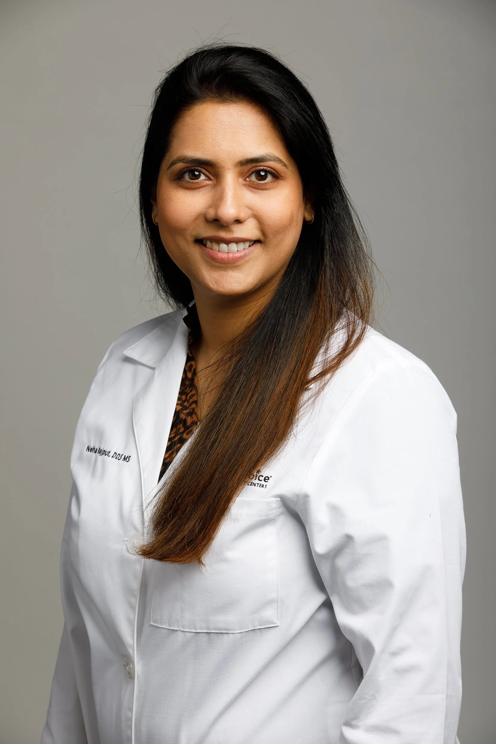A woman with long dark hair wearing a white lab coat, smiling against a gray background. Photo by headshot photographer Tom Krueger, Milwaukee, Wisconsin.