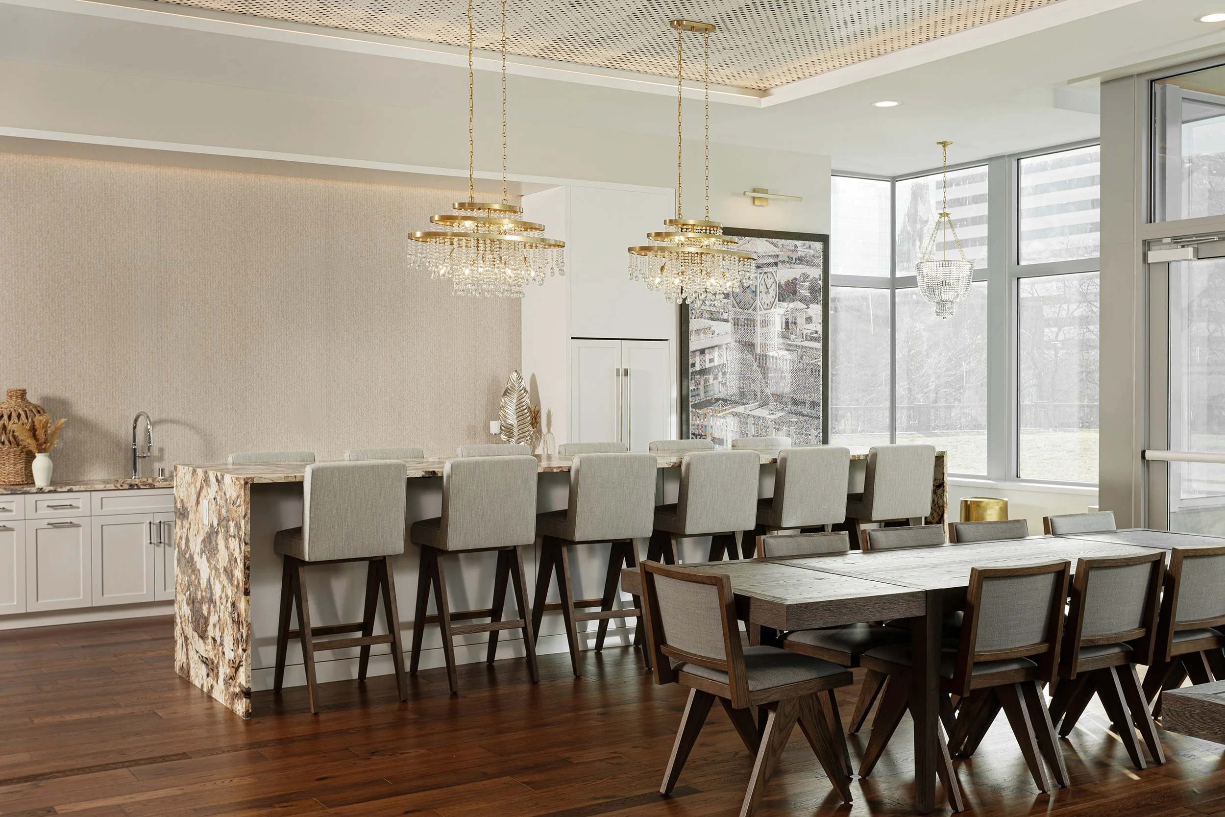 Modern dining room with a long wooden table, beige chairs, and chandeliers. Large windows let in natural light, and a kitchen counter with a patterned stone surface is visible.
