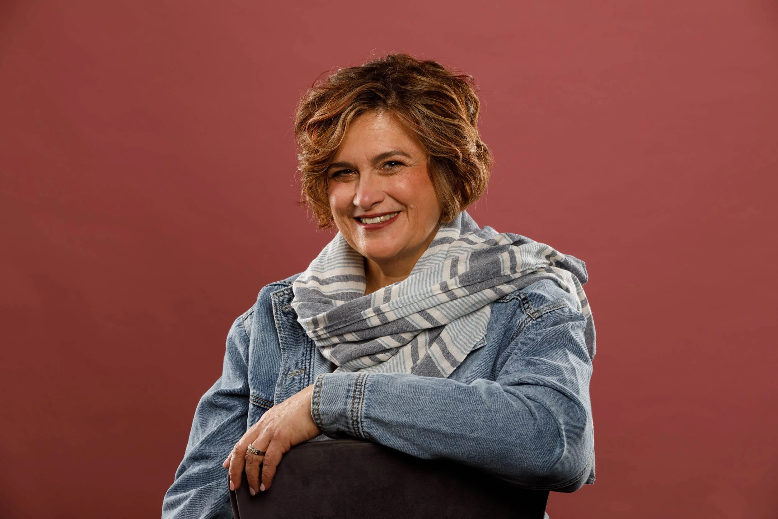 A woman with short, wavy light brown hair, smiling with eyes squinted, wearing a striped scarf and denim jacket, sitting with arms crossed against a plain reddish-brown background.