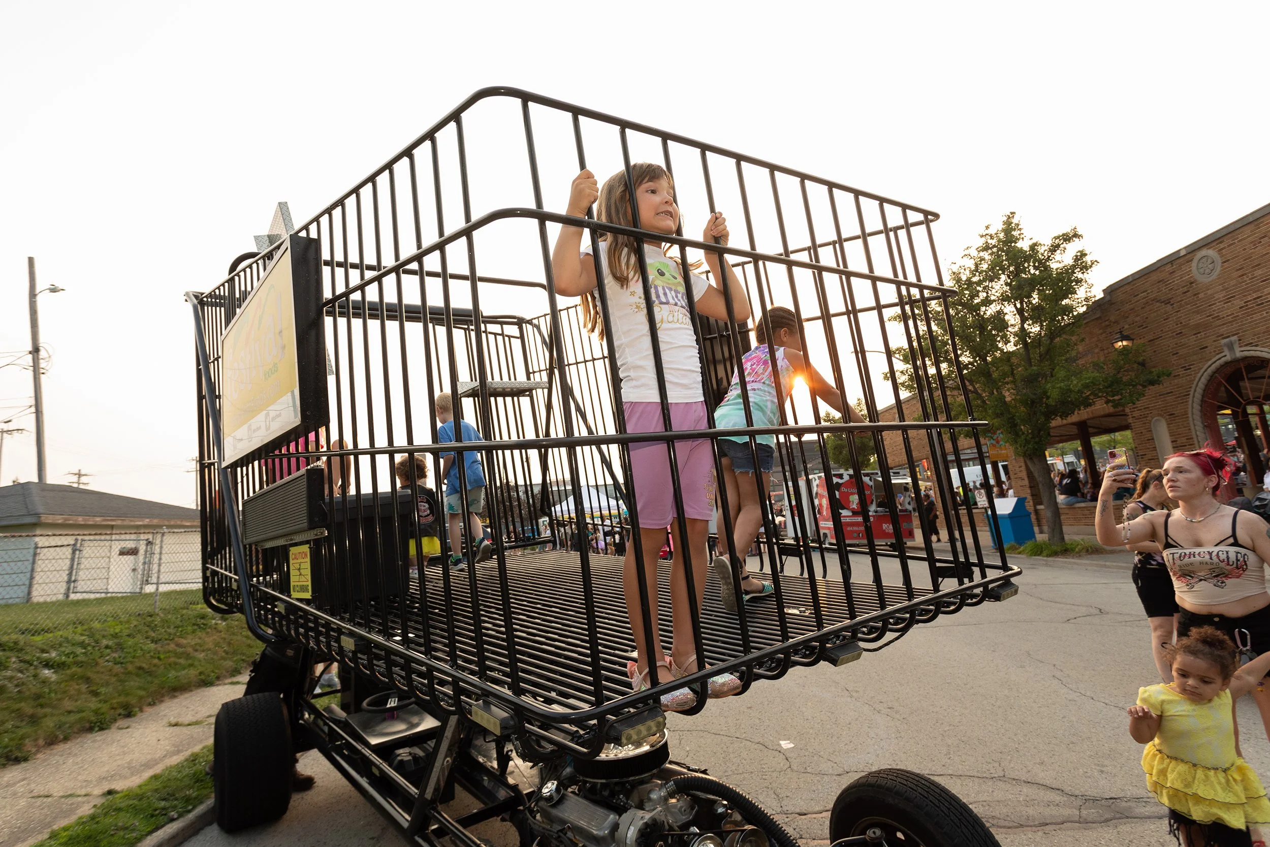 A young girl stands inside a moving black metal cage, holding onto the bars and looking to her right with a concerned expression. Other children and adults are visible around her, with some on the ground and others nearby, at an outdoor event in the 